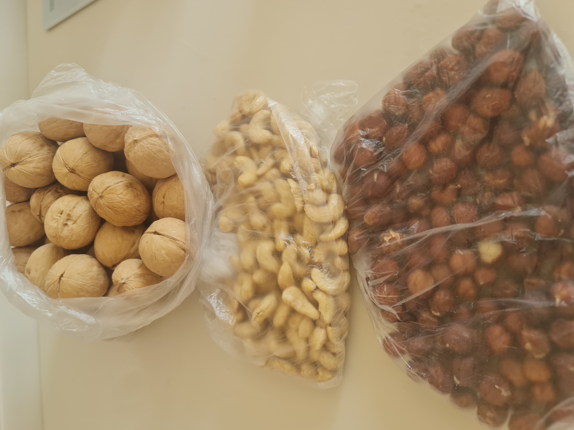 The image shows three plastic bags filled with nuts on a light-colored surface. The top bag contains whole walnuts in their shells. The middle bag is filled with a light-colored, curved nut, likely cashews. The bottom bag contains smaller, reddish-brown nuts, possibly hazelnuts or peanuts. The lighting is overhead, casting faint shadows. The image is an indoor still life and does not provide any information about the city of Angarsk, Russia, or any people or actions.