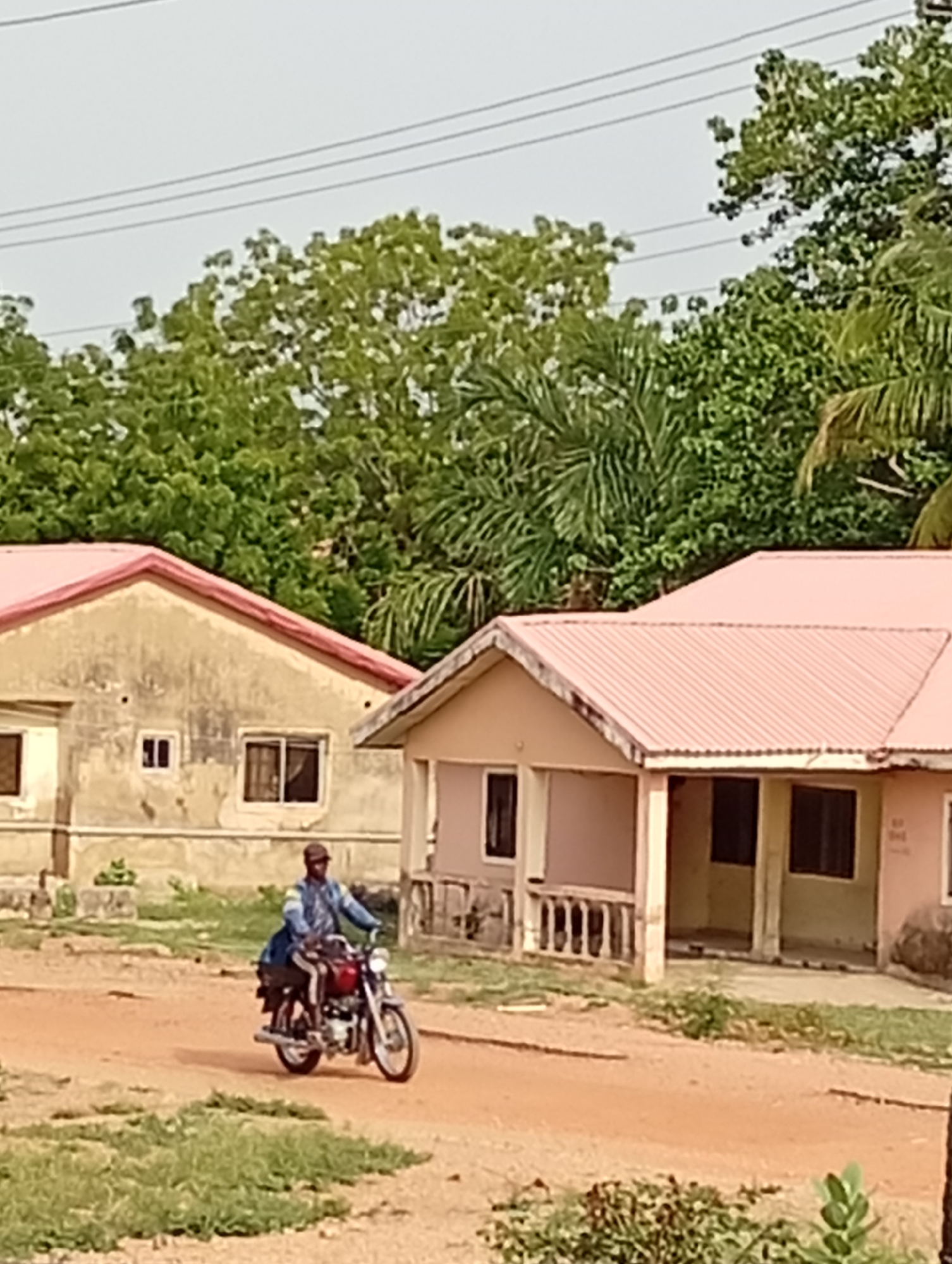 A man is riding a red motorcycle on a dirt road in Adankolo, Nigeria. Two single-story houses with reddish-pink roofs are on either side of the road. The house on the left is beige with a weathered facade, while the house on the right is a light tan color and has a small porch with a railing. Trees and vegetation are visible in the background. Power lines stretch across the sky above.