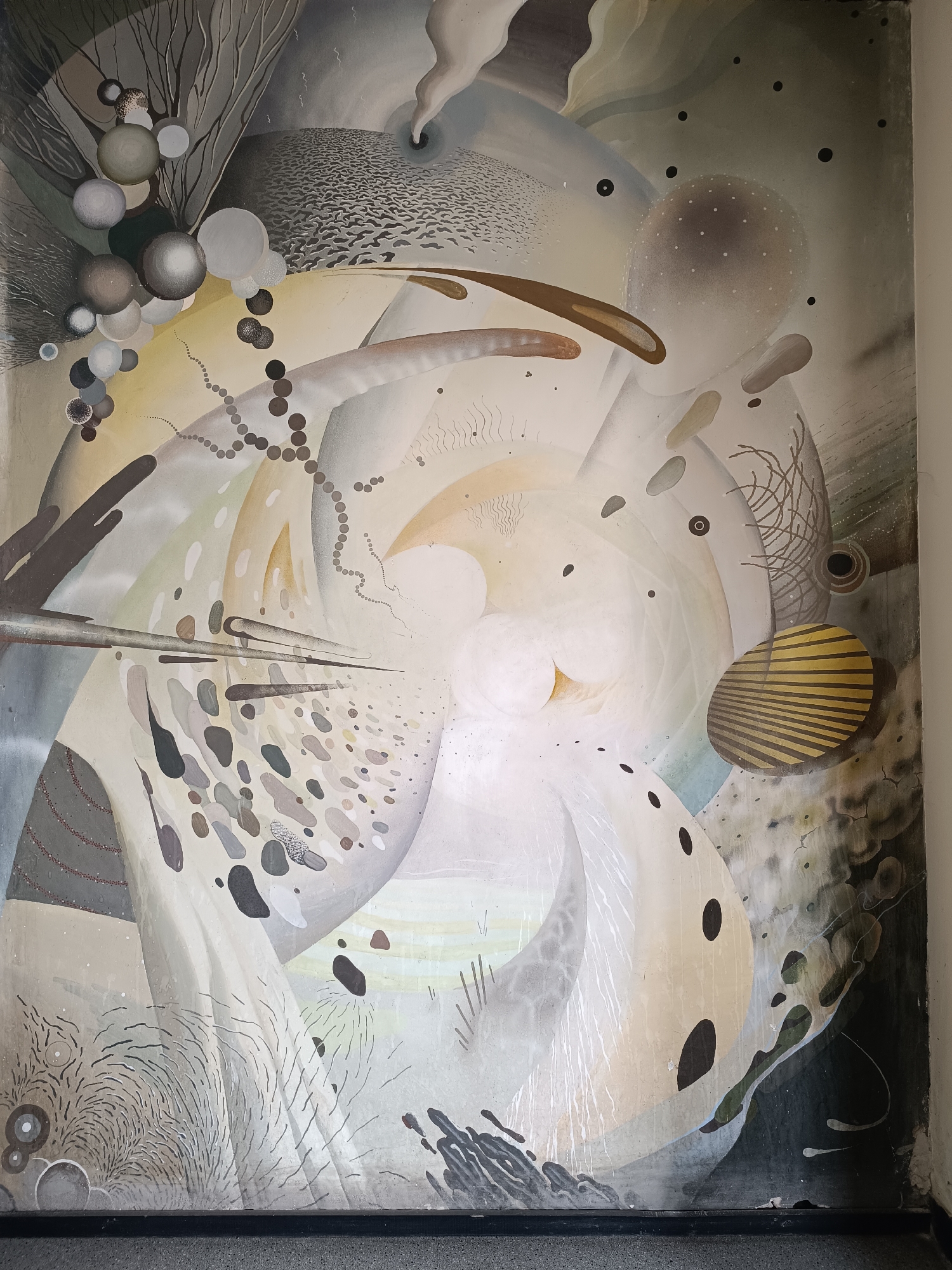 The image shows an abstract painting on a wall in a public space. The painting features a swirling composition of various shapes and textures, including spheres, lines, dots, and cloud-like forms. The color palette is dominated by muted tones of gray, white, brown, and beige. The bottom edge of the painting meets a dark-colored baseboard or floor. The painting is damaged with paint streaks and discoloration. The location is Sofia, Bulgaria.