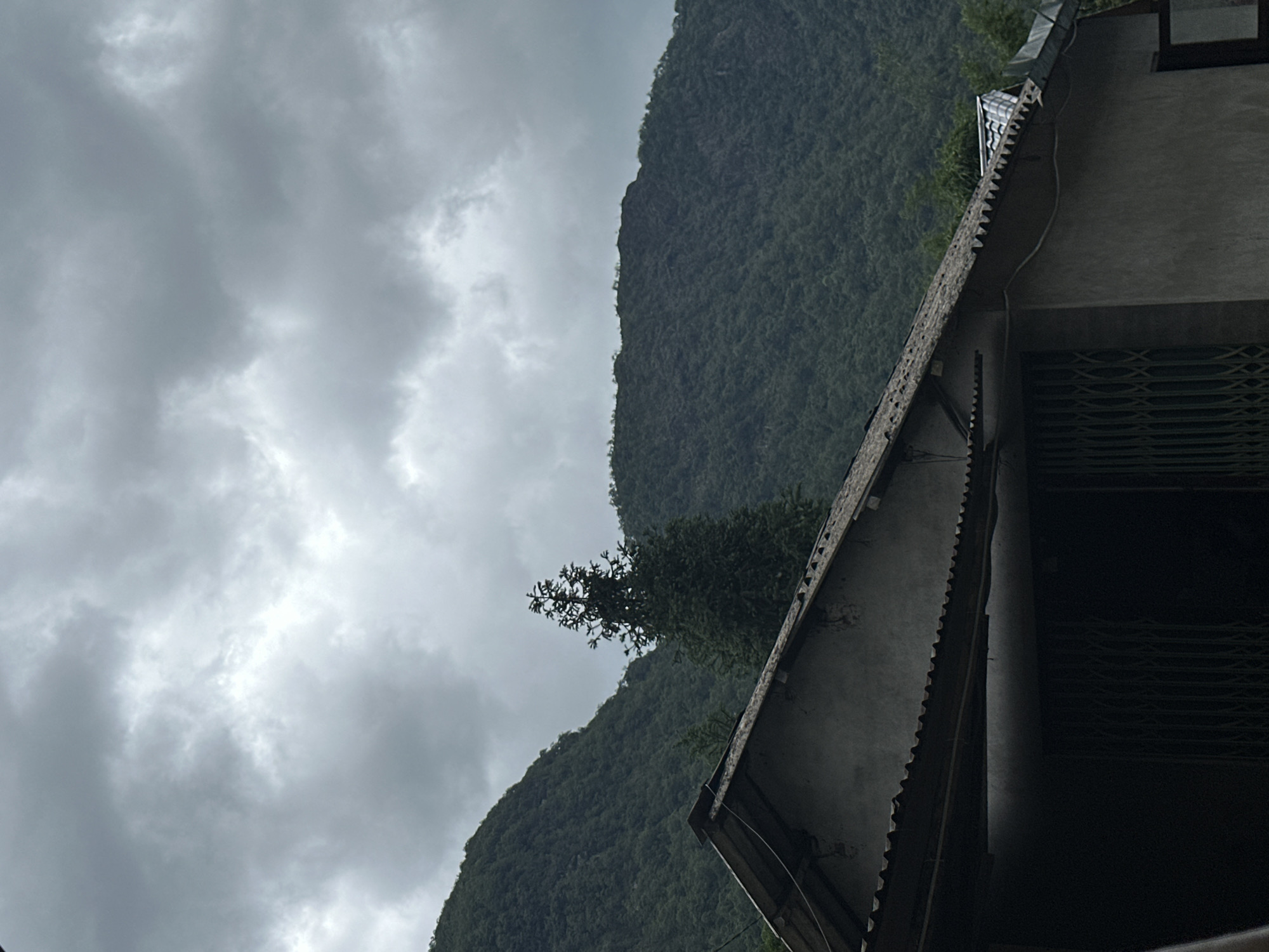 The image shows a landscape in Mường Hoa, Vietnam, featuring a cloudy sky and lush green mountains. In the foreground, part of a building is visible with a corrugated iron roof. The focus is on the natural environment, where the mountain slopes are densely covered with trees and vegetation.
