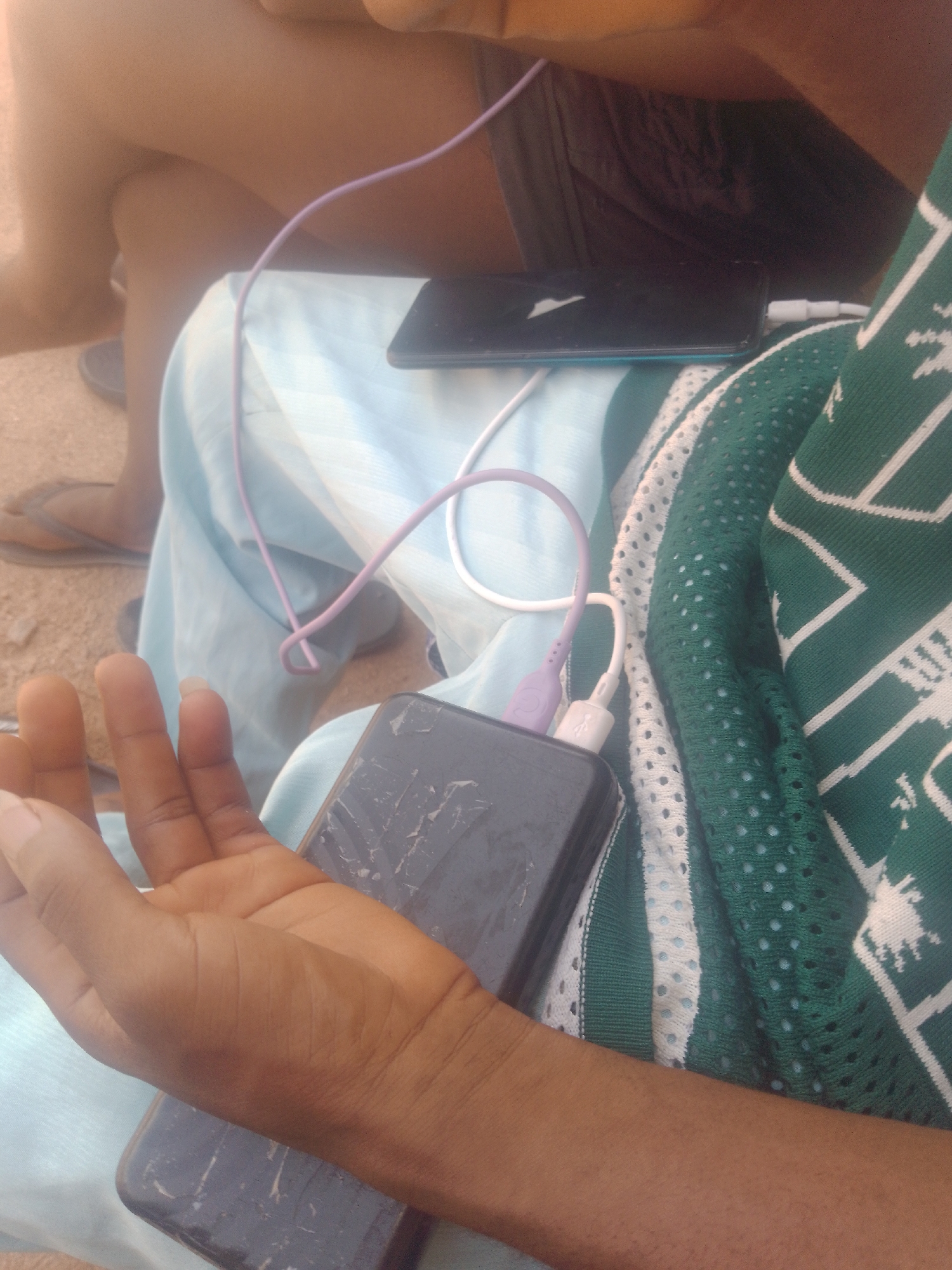 The image shows a close-up view of a person's hands and arms, along with some electronic devices and clothing. Two smartphones are visible, both appearing to be charging. One is dark-colored and has a cracked screen, resting on a light blue fabric. The other, also dark, is partially obscured and connected by white and purple charging cables to what appears to be a power bank. The person's skin tone is dark, and their fingernails are visible. They are wearing a green and white patterned garment with a mesh-like texture. The background is out of focus but suggests an outdoor setting with sandy or dusty ground and another person's legs and feet wearing flip-flops. The lighting indicates it is daytime. The overall scene suggests a casual or informal moment, possibly related to technology use or travel. No visible text is present in the image. The location context is Jauro Yinu, Nigeria.