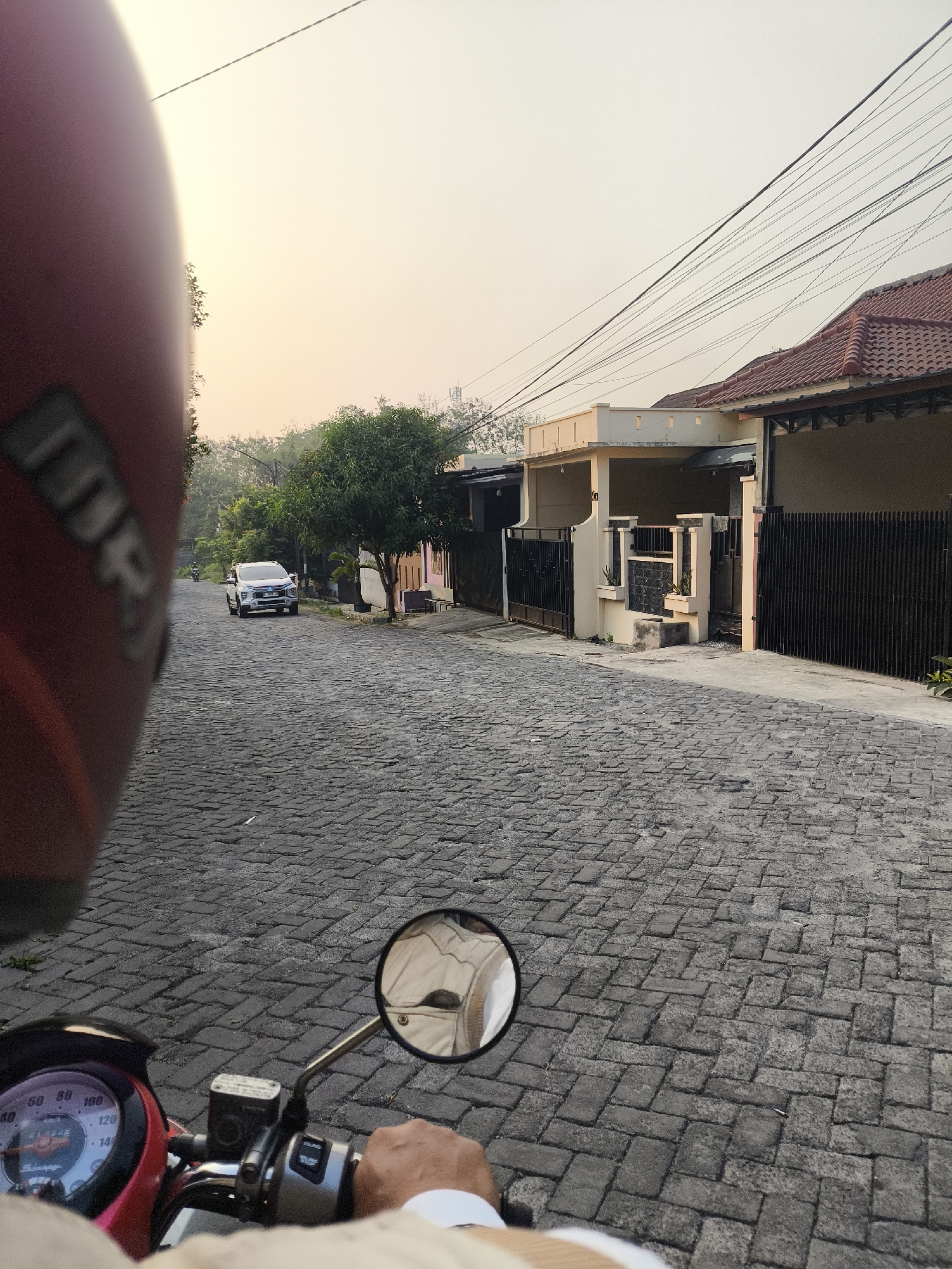 The image depicts a view from the perspective of someone riding a red scooter on a cobblestone street in Cilegon, Indonesia, during what appears to be early morning or late afternoon. The sky is a pale orange, suggesting sunrise or sunset. In the foreground, the rider's red helmet is partially visible on the left, and their hand is on the scooter's handlebars, with a circular mirror reflecting the rider's upper body. The scooter's dashboard is also visible, showing a speedometer. The street is paved with dark gray cobblestones arranged in a herringbone pattern. A white SUV is parked on the right side of the street, and on the left side, a smaller motorbike is visible in the distance. Houses with tiled roofs and black metal fences line the street. Lush green trees and foliage are present, particularly on the left side of the street and near the houses. Electrical wires are strung across the sky.