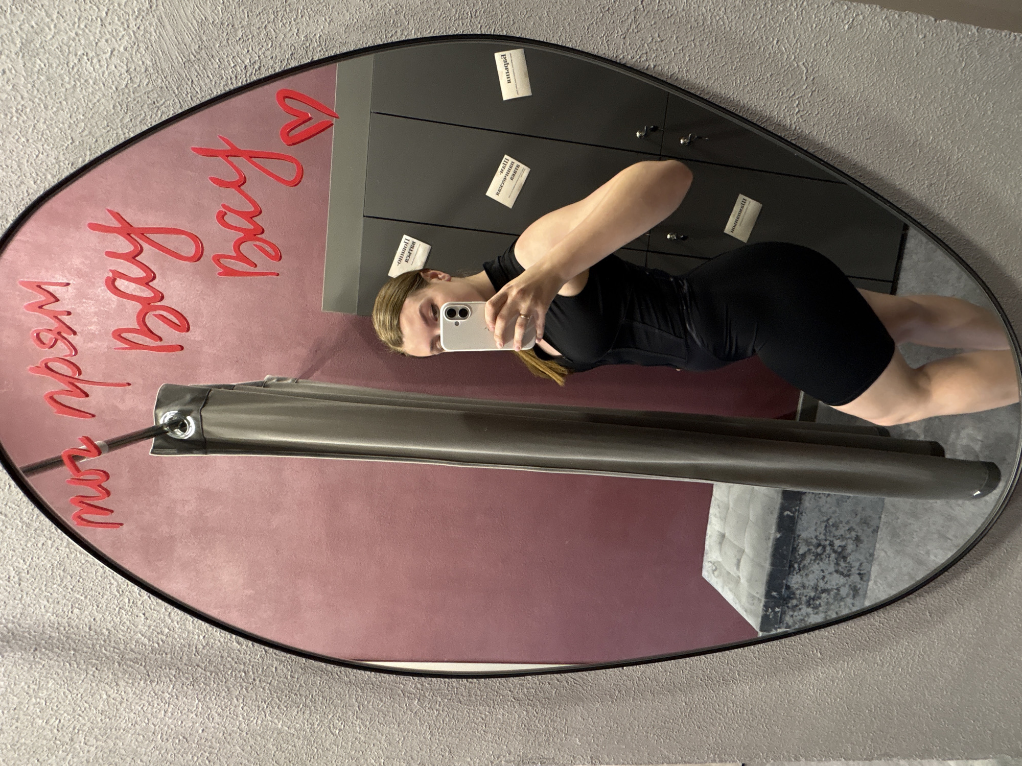 A mirror reflects a woman in a black athletic outfit taking a selfie with a smartphone. The reflection includes lockers in the background. The mirror is mounted on a wall, featuring red decorative text that reads "изряд bay bay <3". The scene is located in Izhevsk, Russia, based on the text.