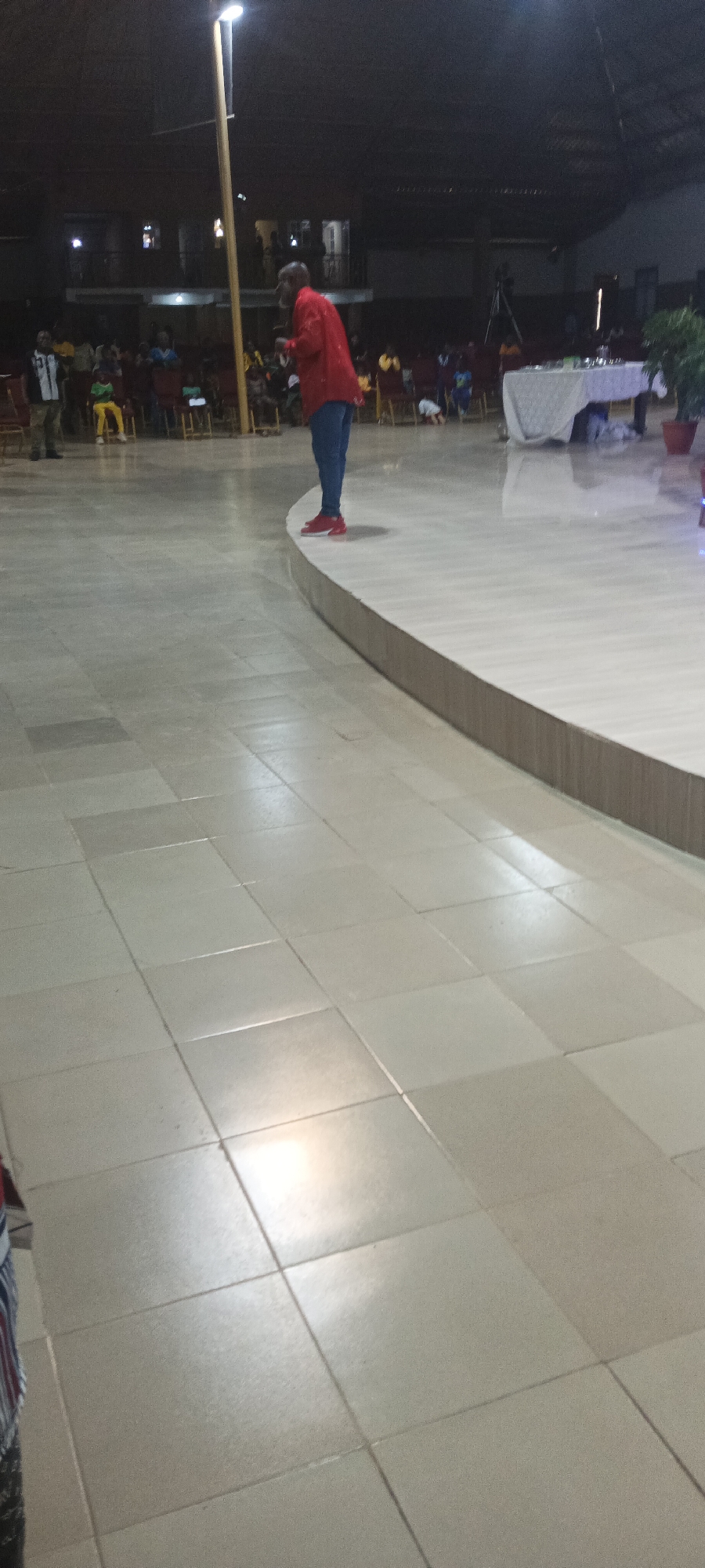 The image shows an interior space, possibly a church or large hall, located in Rukuba Road, Nigeria. A man in a red shirt and blue jeans stands on a raised platform. In the background, rows of chairs are filled with people. There is a table with a white tablecloth. A tall lighting fixture is positioned to the left of the man. The floor is tiled.