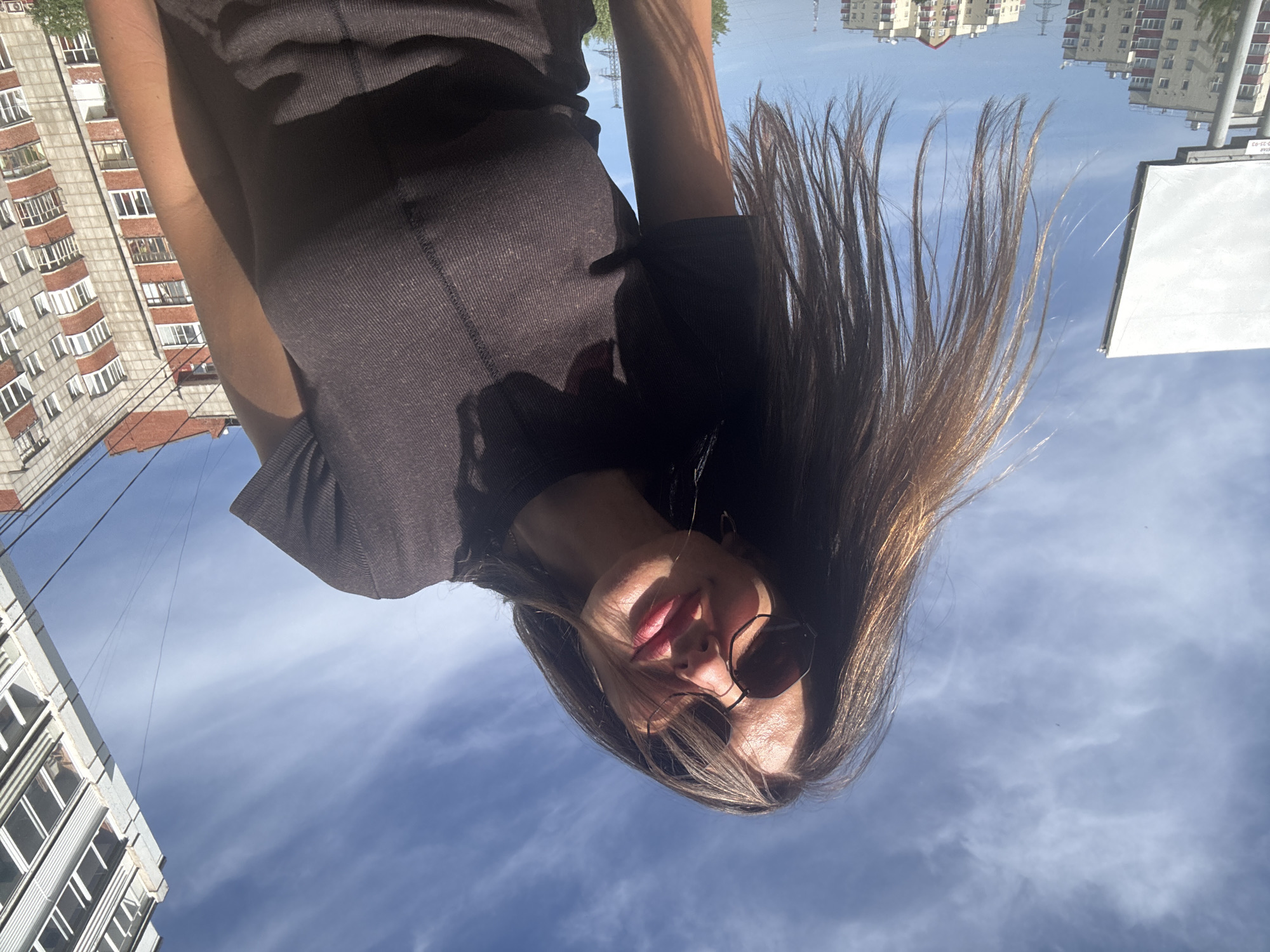 A woman in sunglasses and a dark top is looking at the camera. Her long brown hair is partially covering her face. The background is the sky with a few clouds and buildings. The location is Chaikovsky, Russia.