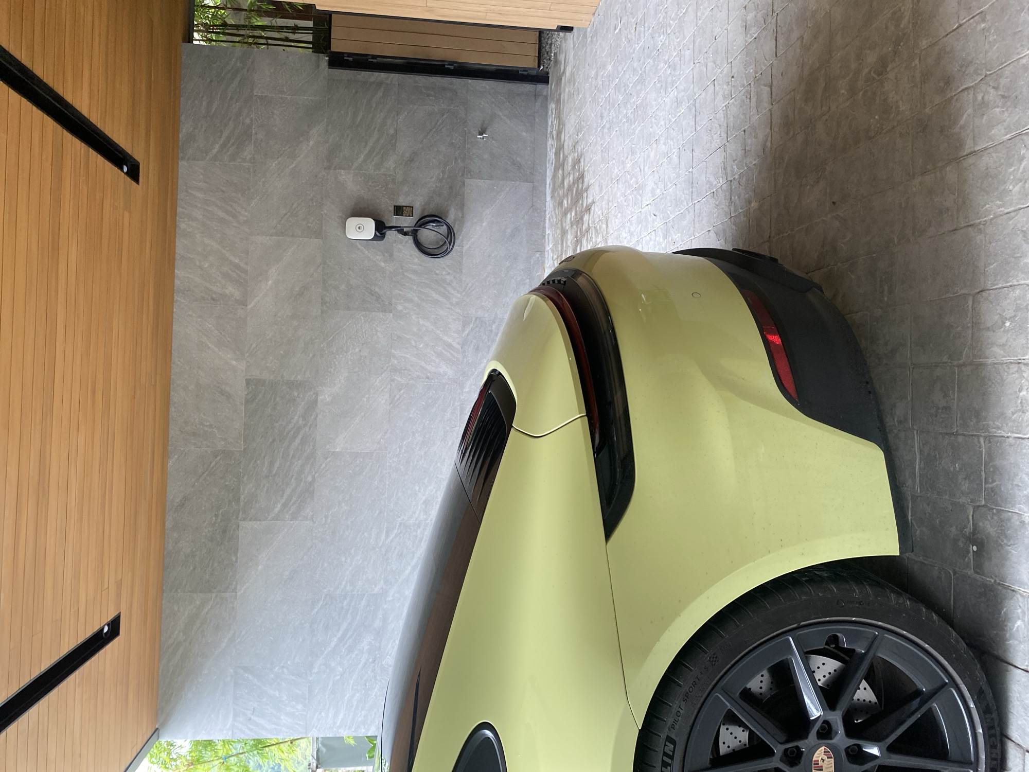 The image shows the rear of a yellow Porsche Taycan parked in what appears to be a garage or covered parking area. The car is positioned on the right side of the frame. A charging station is visible on the gray tiled wall behind the car. The garage has a wooden paneled wall on the left and a tiled floor. The location is likely in a private residence or building in Choeng Thale, Thailand, based on similar architectural styles and residential structures found in that area. The focus is on the car and its charging environment.