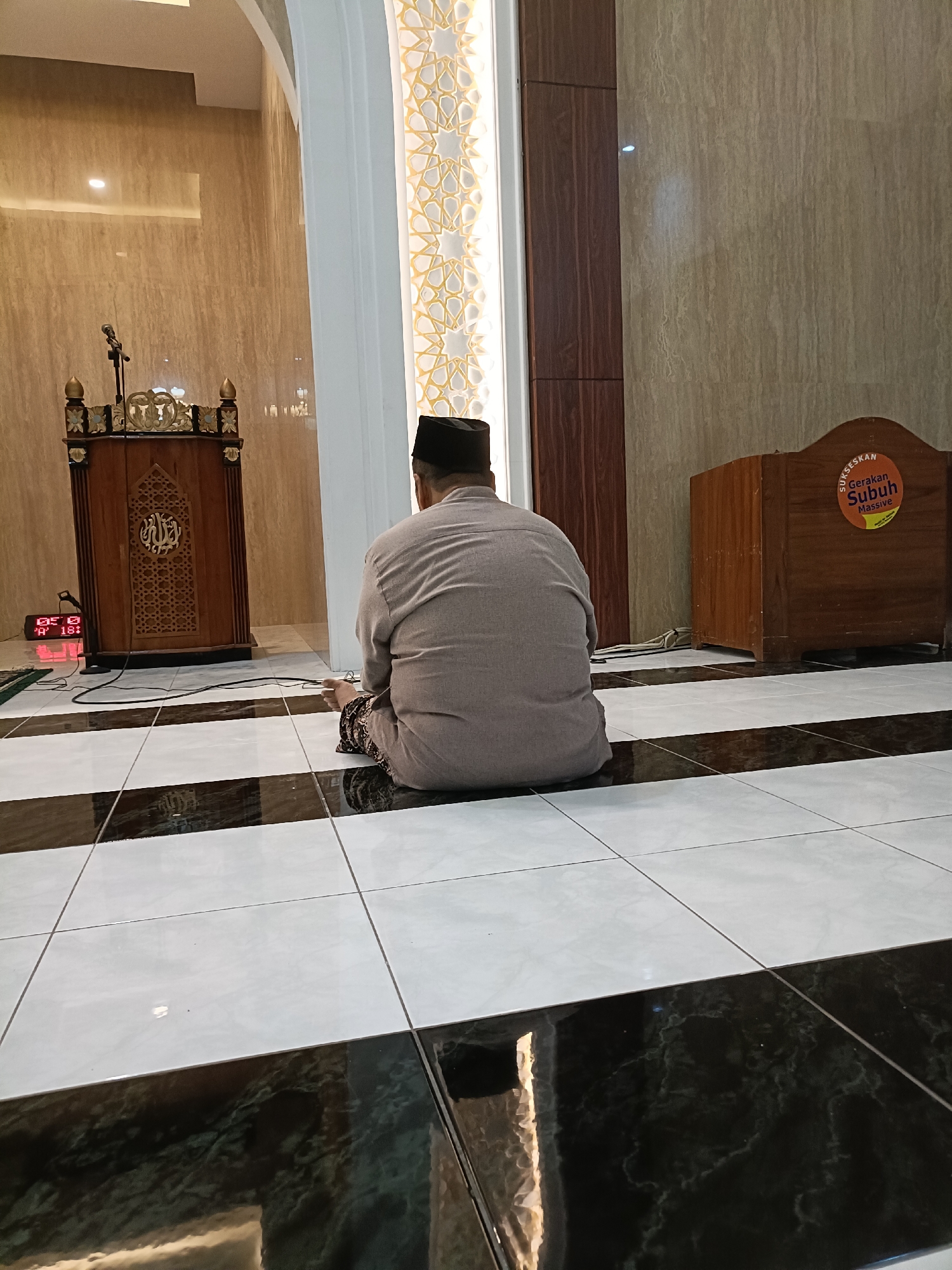 A man wearing a taqiyah and a grey shirt sits on the floor facing a wooden pulpit in a mosque. The mosque has marble walls and a patterned tiled floor. A digital clock shows "05:18".