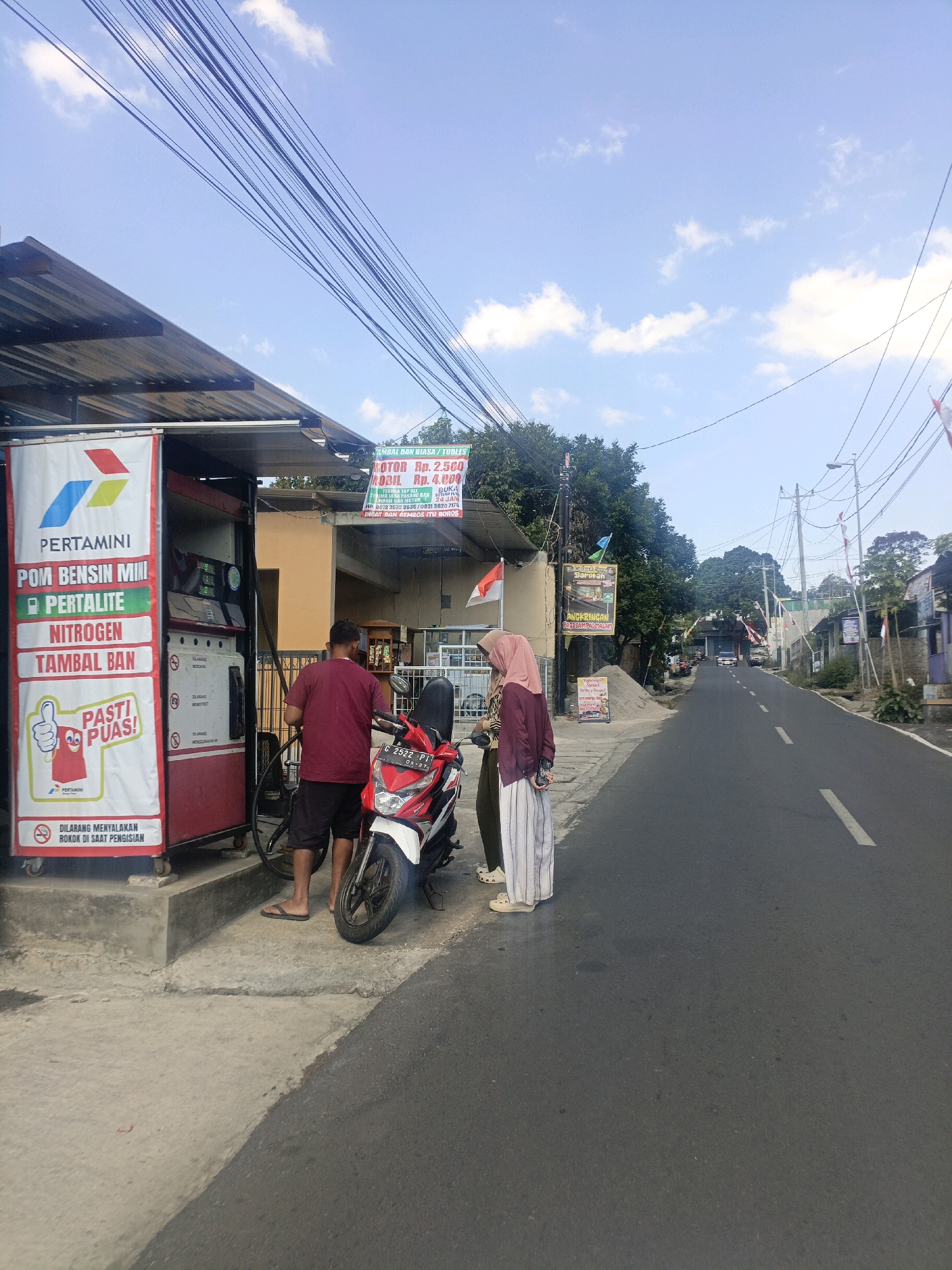 A daytime, eye-level, outdoor, medium shot shows a Pertamina gas station on a street in Salatiga, Indonesia. The gas pump is red and white with a Pertamina logo. A red and white motorcycle is parked in front of it. A man in a maroon shirt and black shorts is standing by the motorcycle, holding a fuel nozzle. A woman in a hijab and a long-sleeved purple shirt is standing next to him. The street is paved and lined with trees and buildings. Overhead, numerous electrical wires crisscross the sky, which is partly cloudy. A sign above the gas station reads "TAMBAL BAN BIASA / TUBLES" and lists prices for motorcycle and car tire repairs. Another sign below the gas pump displays a thumbs-up icon and the text "PASTI PUAS!" and "DILARANG MENYALAKAN ROKOK DI SAAT PENGISIAN."