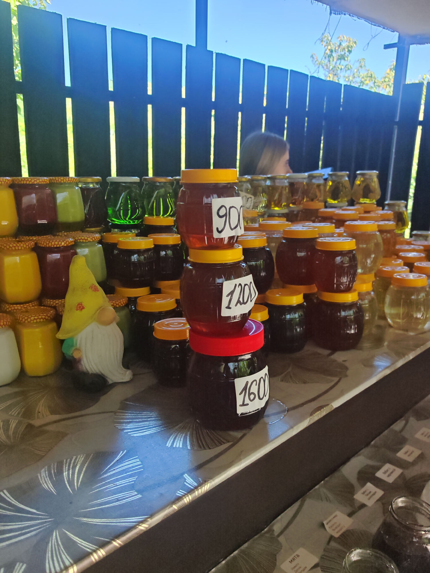 In Varvarovka, Russia, a market stall displays a variety of jars filled with honey and other products. The jars are predominantly glass with yellow or red lids and are arranged on a patterned tablecloth. Some jars have handwritten price tags indicating "900," "1200," and "1600." A small, ceramic gnome figurine with a yellow hat is positioned amongst the jars. In the background, a person with blonde hair is partially visible, and a dark wooden fence with vertical slats is present. The environment appears to be outdoors, with sunlight casting shadows and illuminating the scene.