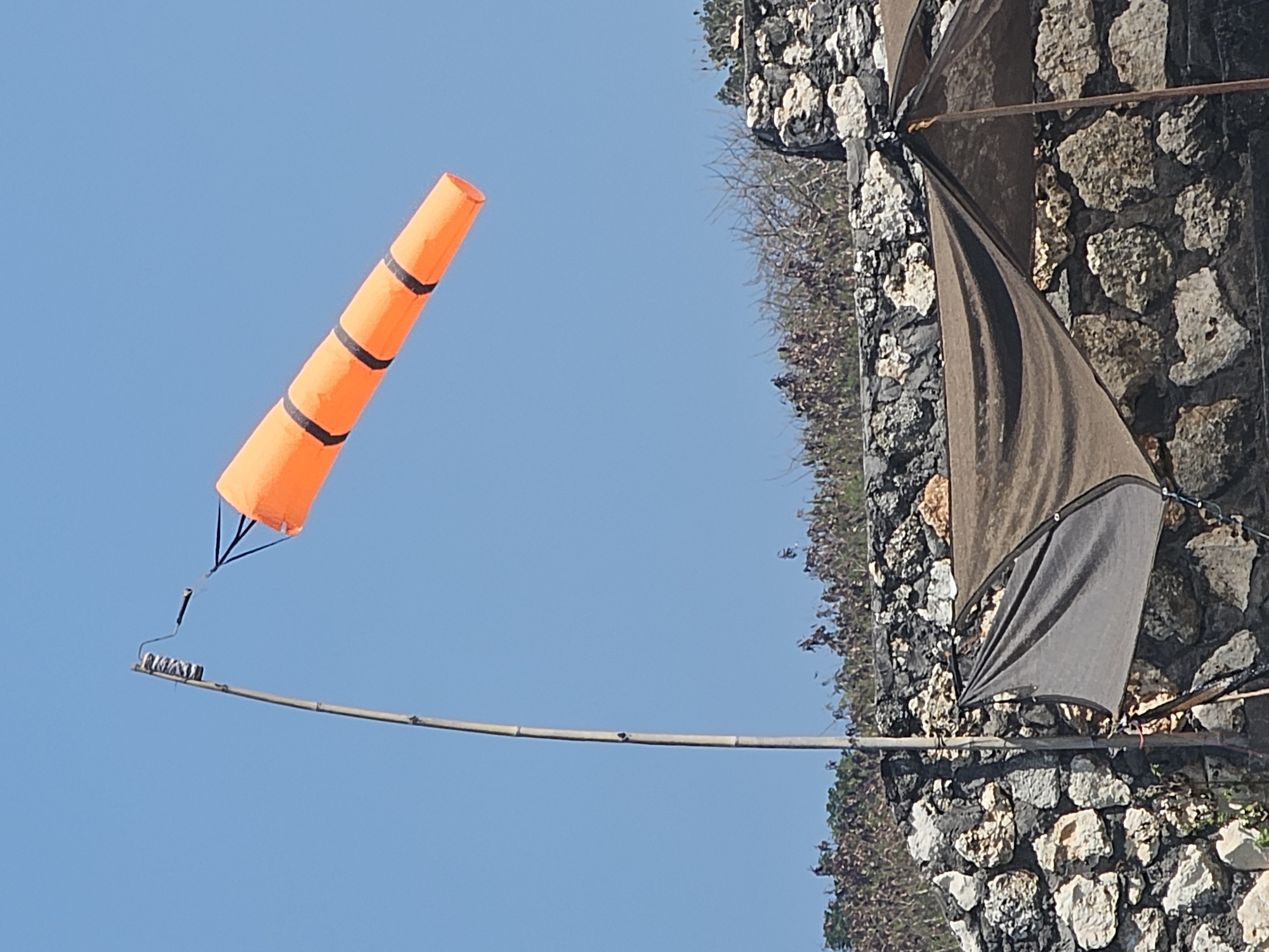 An orange windsock with black stripes is attached to a bamboo pole and is blowing in the wind against a clear blue sky. Below the windsock and pole, there is a stone wall. On top of the stone wall, there are dry bushes and some trees. Partially visible in the foreground, below the stone wall, are two shades: one dark gray and one brown. The scene appears to be outdoors, likely in a coastal area given the dry vegetation and stone construction. Kuta Selatan is a sub-district in Bali, Indonesia, known for its beaches and resorts, which could explain the presence of a windsock.