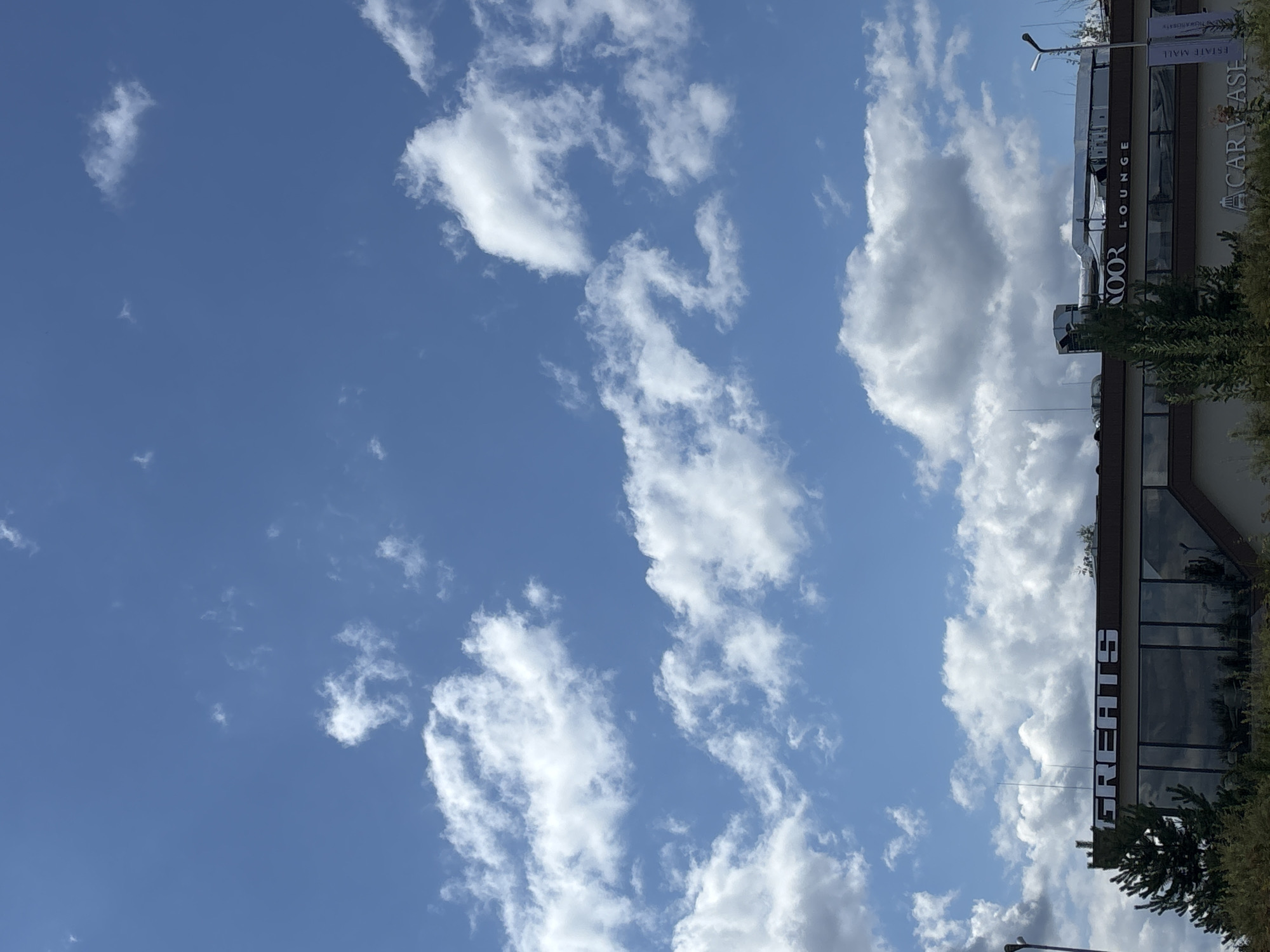 The image captures a building with signage that reads "GREATS" in large white letters and "LOUNGE" in smaller white letters. The building appears to be a commercial establishment. Above the building, a vast blue sky is filled with scattered white clouds, varying in size and density. To the lower right of the building, a banner displaying "CAR WASH" is visible. Trees and foliage surround the base of the building, suggesting an outdoor setting. The scene is consistent with a location in Millennium Park, Russia.