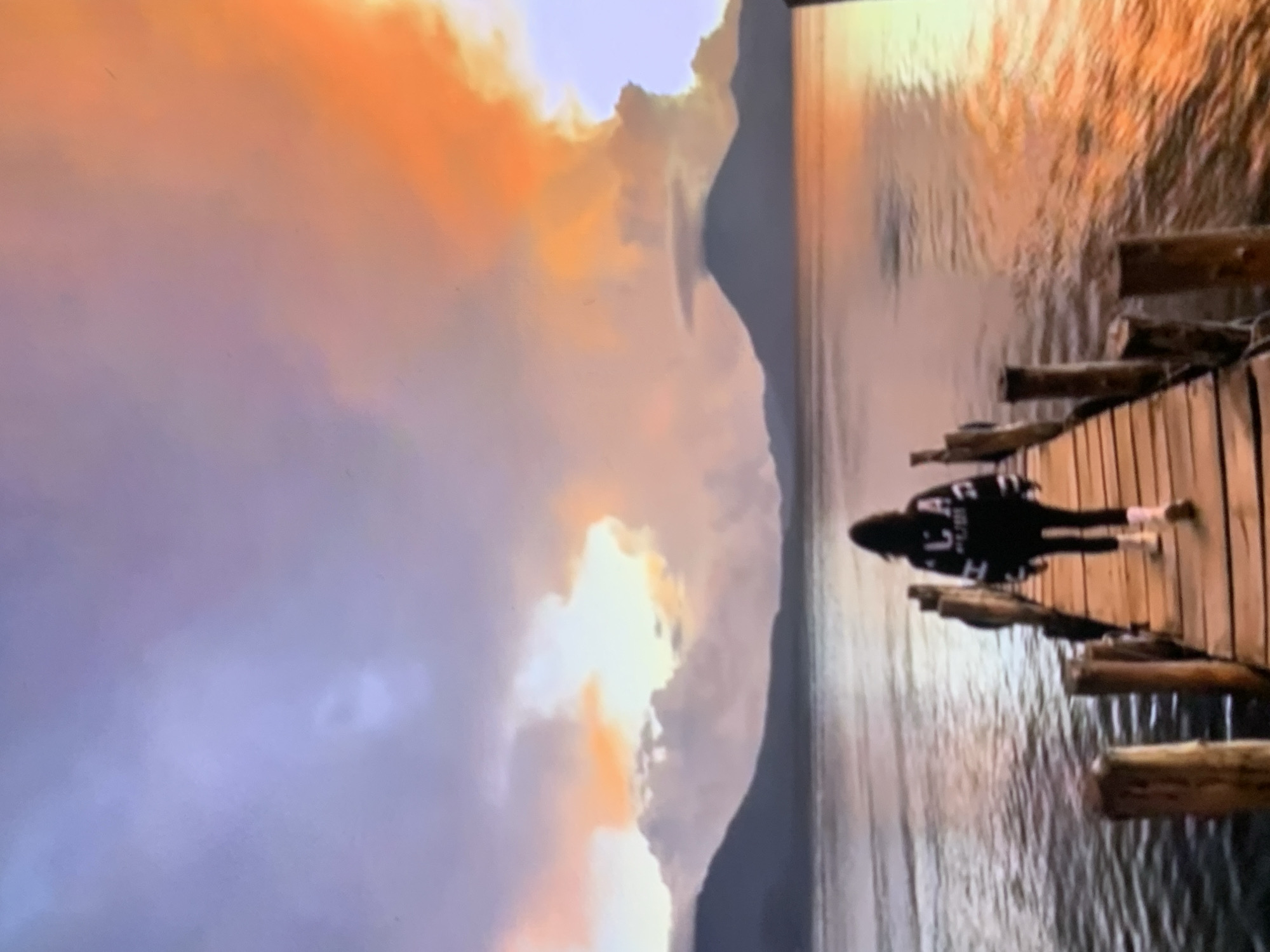 A lone individual walks down a wooden pier extending into a body of water, towards a dramatic sunset. The sky is ablaze with vibrant colors, ranging from soft purples and blues to intense oranges and yellows, with large, illuminated clouds dominating the upper half of the frame. In the distance, silhouetted mountains form a horizon line across the water. The pier is constructed of weathered wooden planks and supported by rustic wooden posts. The person, seen from behind, is wearing dark clothing with white lettering on the back of their sweater and light-colored boots. The water reflects the colors of the sky, creating a shimmering, colorful surface. The scene evokes a sense of tranquility and natural beauty. There is no discernible connection to Stavropol, Russia.