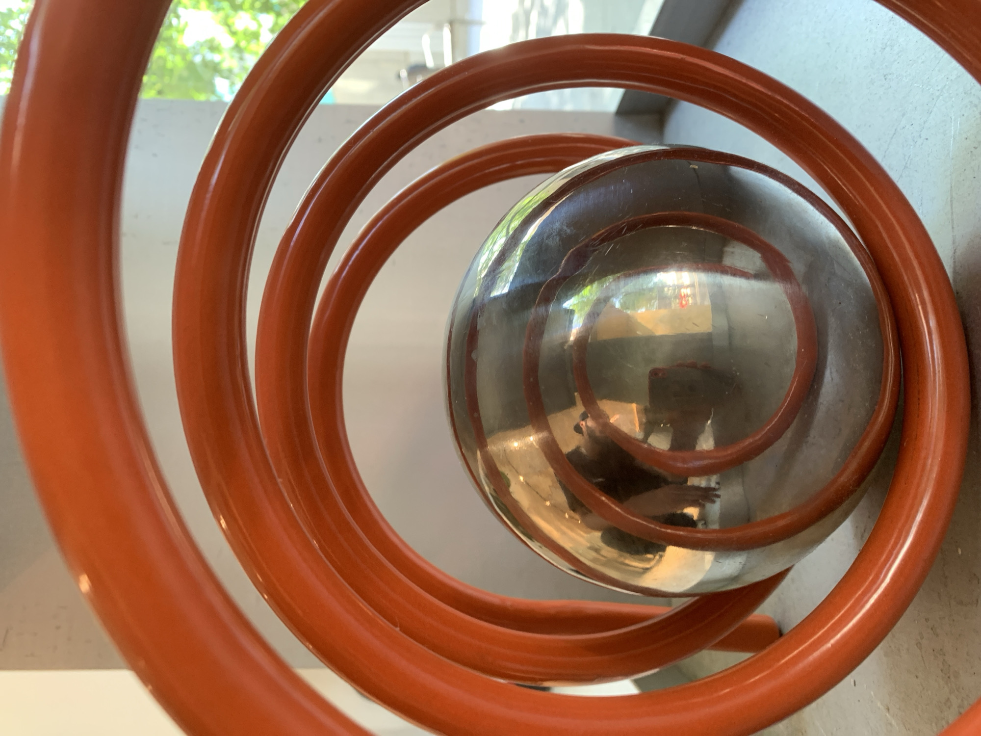 The photograph depicts an abstract sculpture with a large, shiny metal sphere cradled within an orange-colored, spiraling tubular structure. The sculpture appears to be indoors, positioned against a light-colored wall. The metal sphere reflects its surrounding environment, capturing blurred images of people and objects, including a person holding a phone, presumably the photographer. The location is hinted to be Barcelona, Spain, though the image does not provide concrete evidence to verify it.