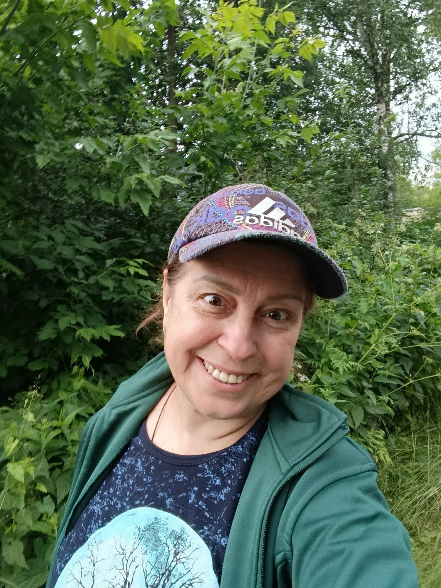 A woman is captured in a selfie outdoors, with lush green foliage forming the backdrop. She is wearing a green zip-up hoodie over a dark blue t-shirt adorned with a light blue graphic of a tree. On her head, she sports a colorful baseball cap with an Adidas logo visible. Her expression is a wide, toothy grin, and her eyes are wide. The scene suggests a casual outing in a natural setting, potentially in the city of Vereya, Russia, given the mention of the location.