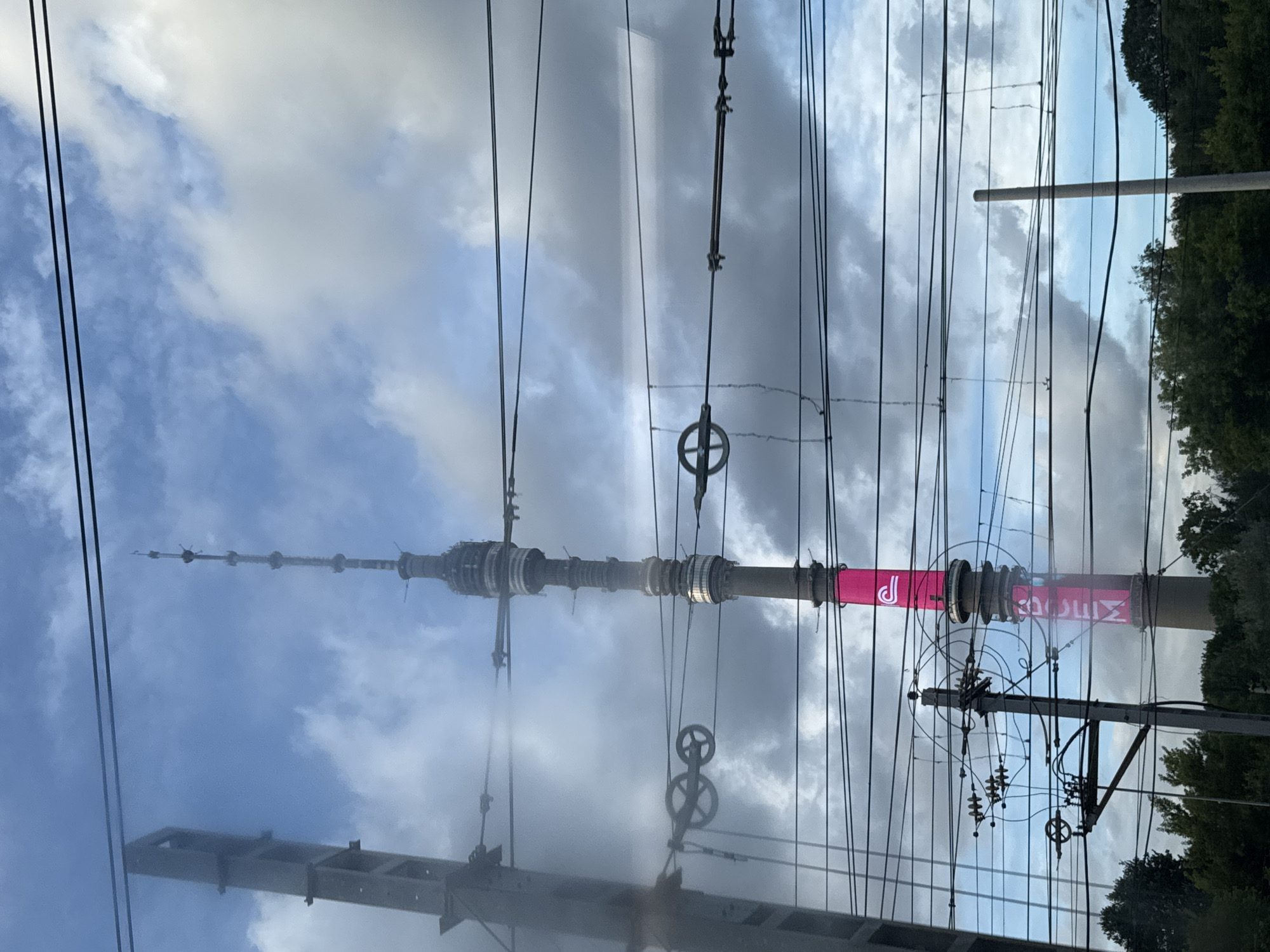 The image captures a view of the Ostankino Tower in Khimki, Russia, seen through a network of overhead power lines and train catenary wires. The tower, a prominent landmark, is partially obscured by the wires, which stretch horizontally across the frame. The tower itself is a tall, slender structure with a distinctive antenna at its peak and multiple levels and platforms. A large, bright pink advertisement is visible on the lower section of the tower, featuring white lettering. To the left of the tower, a tall, grey concrete support structure for the power lines is visible. The sky above is filled with a mix of blue sky and large, dramatic clouds. Trees and foliage are visible at the bottom of the frame, suggesting a location near a wooded area or park. The overall impression is one of urban infrastructure intertwined with a significant architectural element against a cloudy sky.