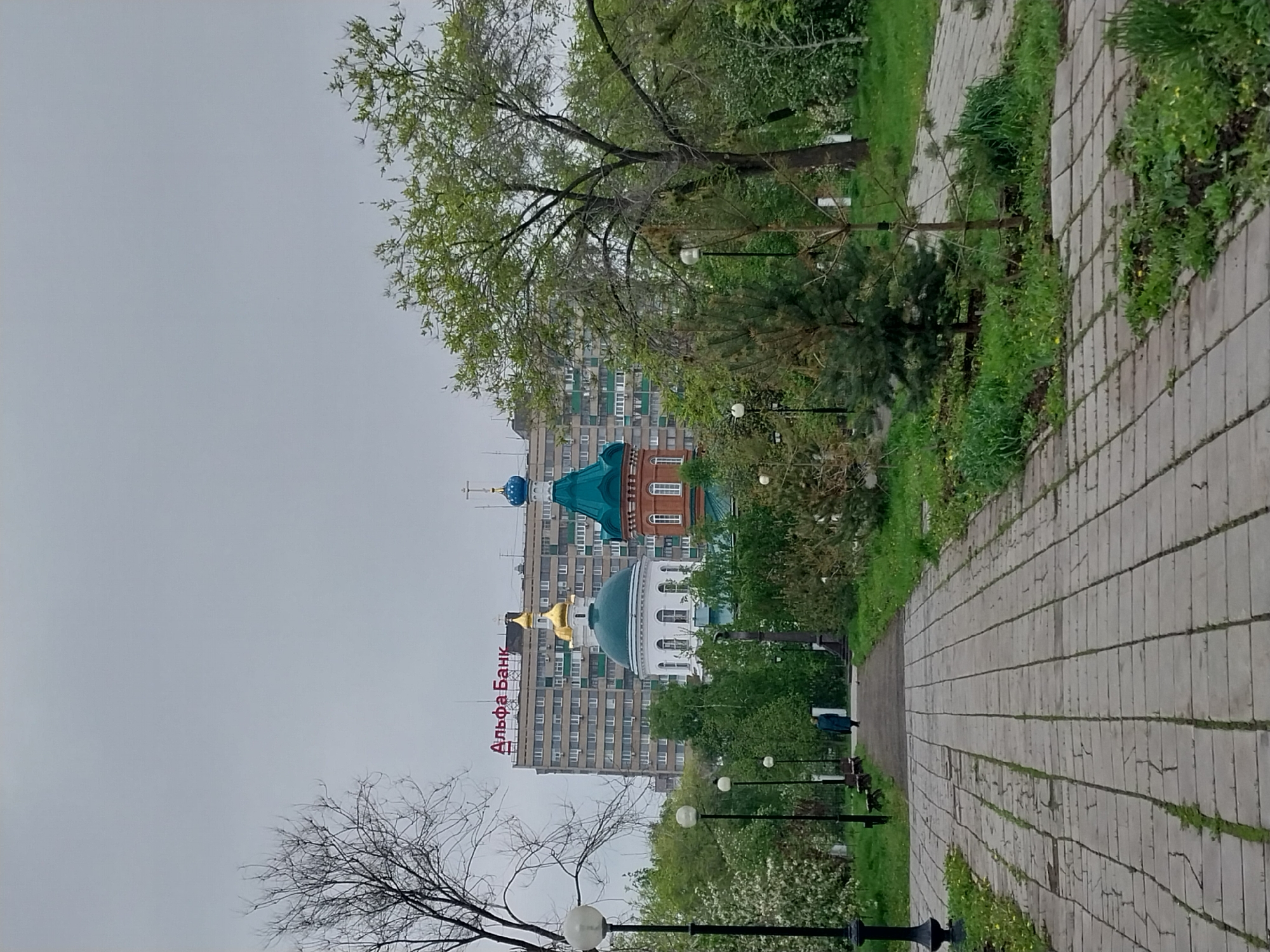 The photograph depicts a park scene in Orenburg, Russia, on an overcast day. A paved pathway stretches into the distance, lined with trees and lamp posts. In the background, buildings are visible, including one with the Russian text "Альфа Банк" (Alfa Bank). The domes of a Russian Orthodox church, colored blue, gold, and green, rise above the buildings. The scene is calm and quiet, with no obvious human activity except for a single figure walking into the distance. The environment appears to be urban, blending into a park setting.