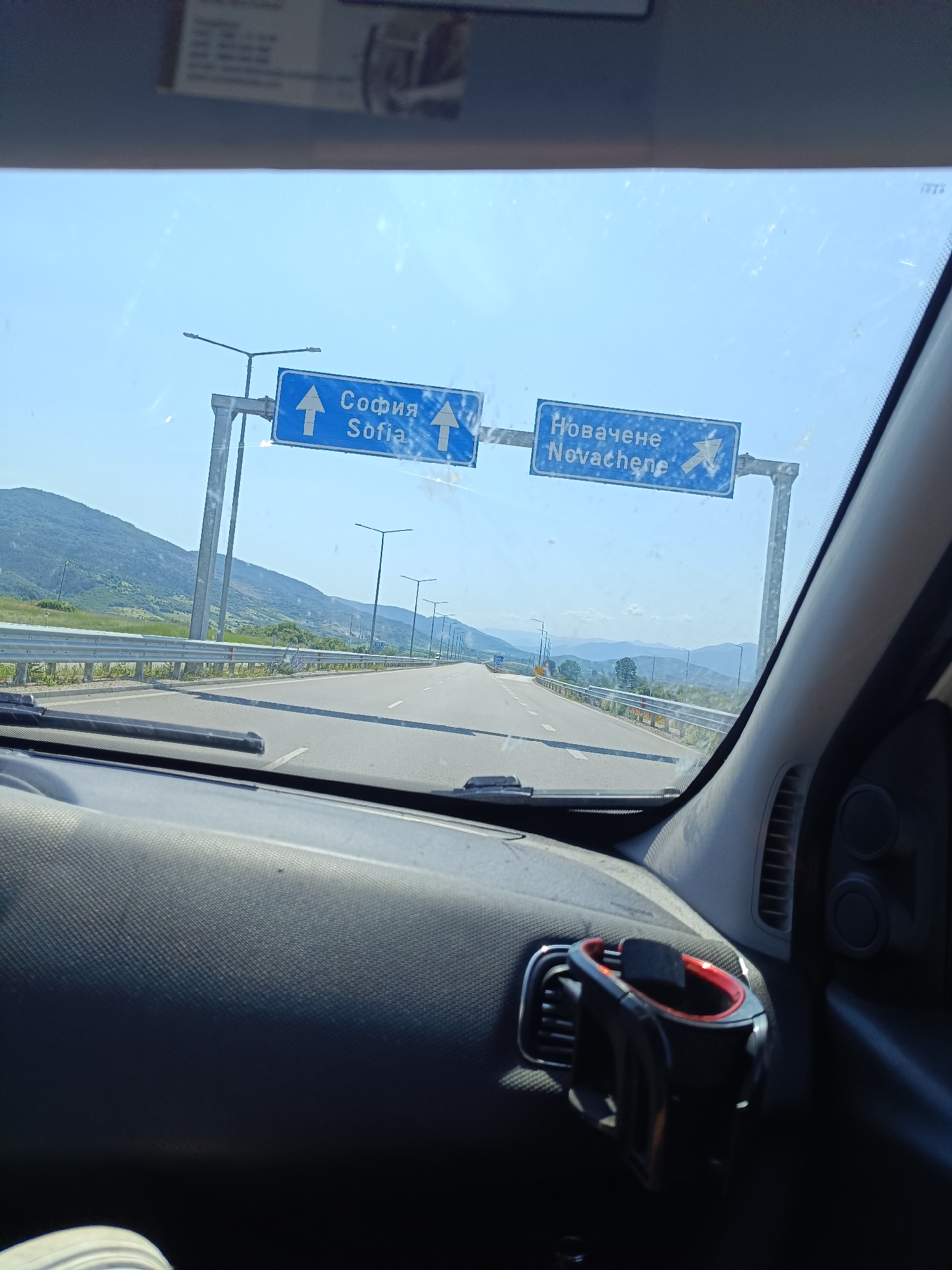 A first-person perspective shot from inside a car shows a highway with directional signs. The sign on the left reads "София Sofia" with an upward arrow. The sign on the right reads "Новачене Novachene" with a rightward arrow. The highway is bordered by a metal guardrail and rolling hills with trees. The sky is clear and blue. The dashboard of the car is visible in the foreground, with a car mount for a phone attached to the air vent.