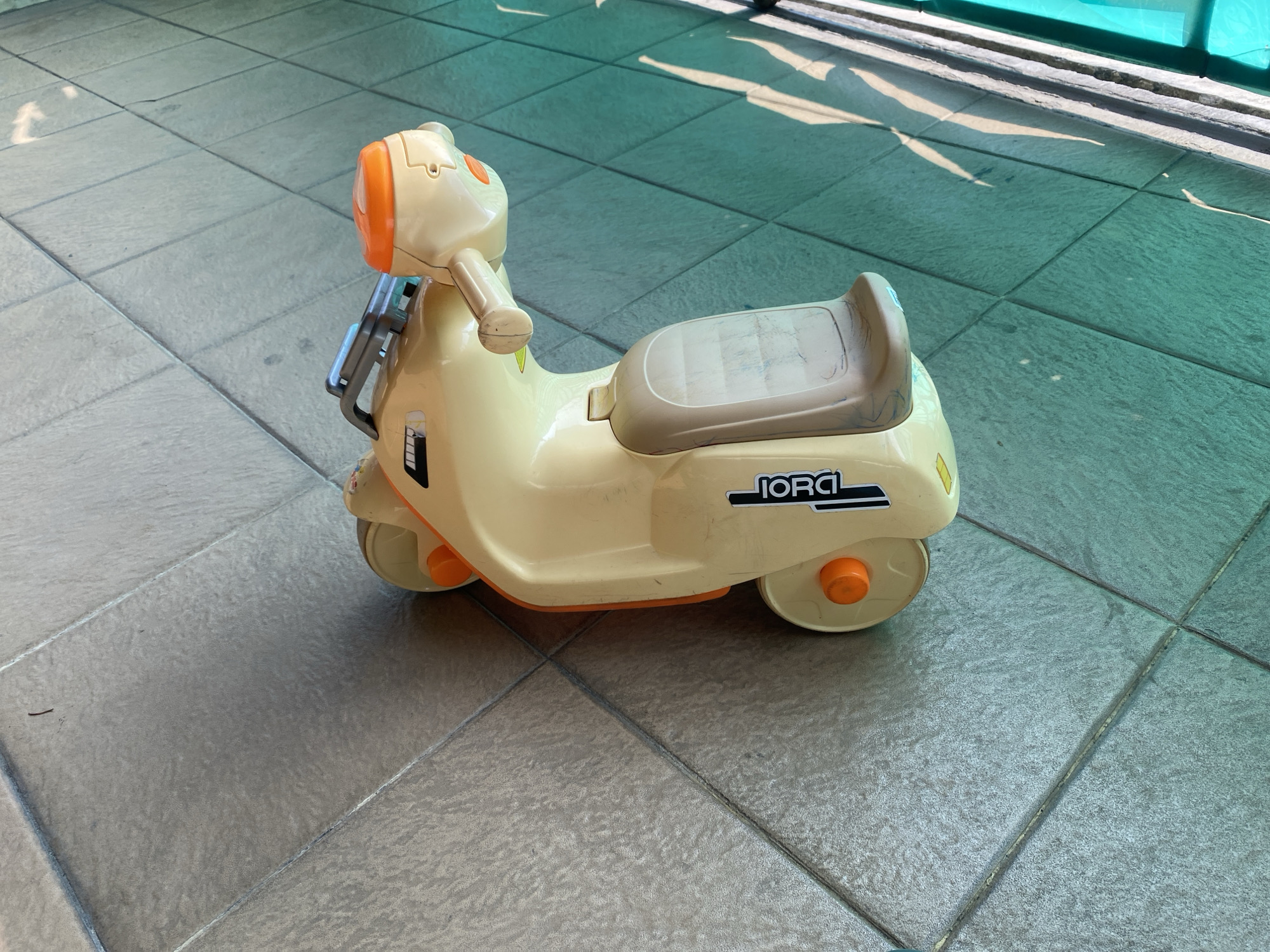 A cream-colored toy scooter with orange accents is parked on a tiled outdoor surface. The scooter has a seat, handlebars, and two visible wheels, with a third wheel possibly hidden from view. A black and white sticker with the word "JORCI" is affixed to the side of the scooter. The tiled surface appears to be part of a walkway or patio, with shadows cast by overhead structures, possibly a green awning or canopy. The lighting suggests it is daytime. This scene could be found in a residential area or a public space in Cilegon, Indonesia.