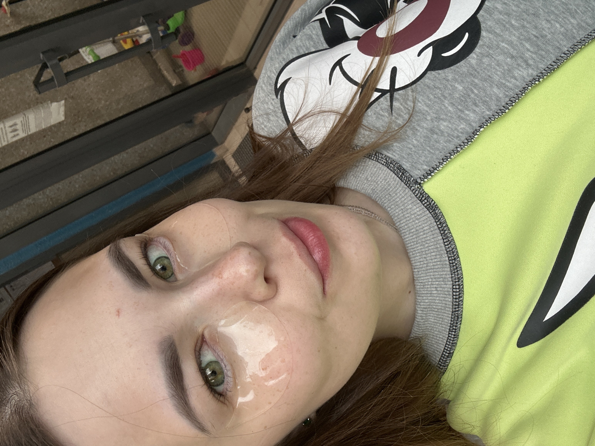 A medium shot captures a young woman with green eyes and brown hair, wearing under-eye patches and a lime green and gray sweatshirt with a character graphic. She is looking towards the camera with a slight smile. The background is blurred, suggesting an indoor setting with glass doors and shelves visible. The lighting is natural and soft. The image is likely a selfie. There is no discernible connection to the city of Perm, Russia.