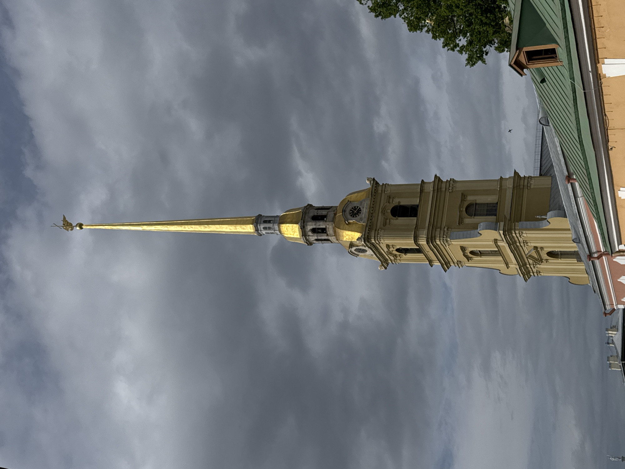 The image presents a striking view of a tall, golden spire and the upper sections of a grand building, captured at an unusual angle due to the photograph being rotated 90 degrees clockwise. The central subject is a slender, luminous golden spire, stretching across the central left portion of the frame. It culminates in an ornate golden weather vane depicting an angelic figure. Below the spire, the building features multi-tiered golden sections, including visible clocks on at least one facade. Further to the right, the structure transitions into a light yellow or cream-colored facade, adorned with classical architectural details such as arched windows and pilasters.
The background of the image is dominated by a heavily overcast sky, filled with thick, dark grey clouds, which occupy the entire upper portion of the rotated frame. To the lower right, details of an adjacent building are visible, including a steep, dark green pitched roof with a small dormer window, and a section of a light orange wall. A patch of green foliage from a tree can be seen in the lower left corner.
The weather appears cloudy and possibly somber, indicative of an overcast daytime. No specific activity is depicted, and no people are visible in the scene. There is no discernible text present on any of the structures. Based on the provided location context, the prominent golden spire and architectural style identify this as the Peter and Paul Cathedral in Saint Petersburg, Russia.