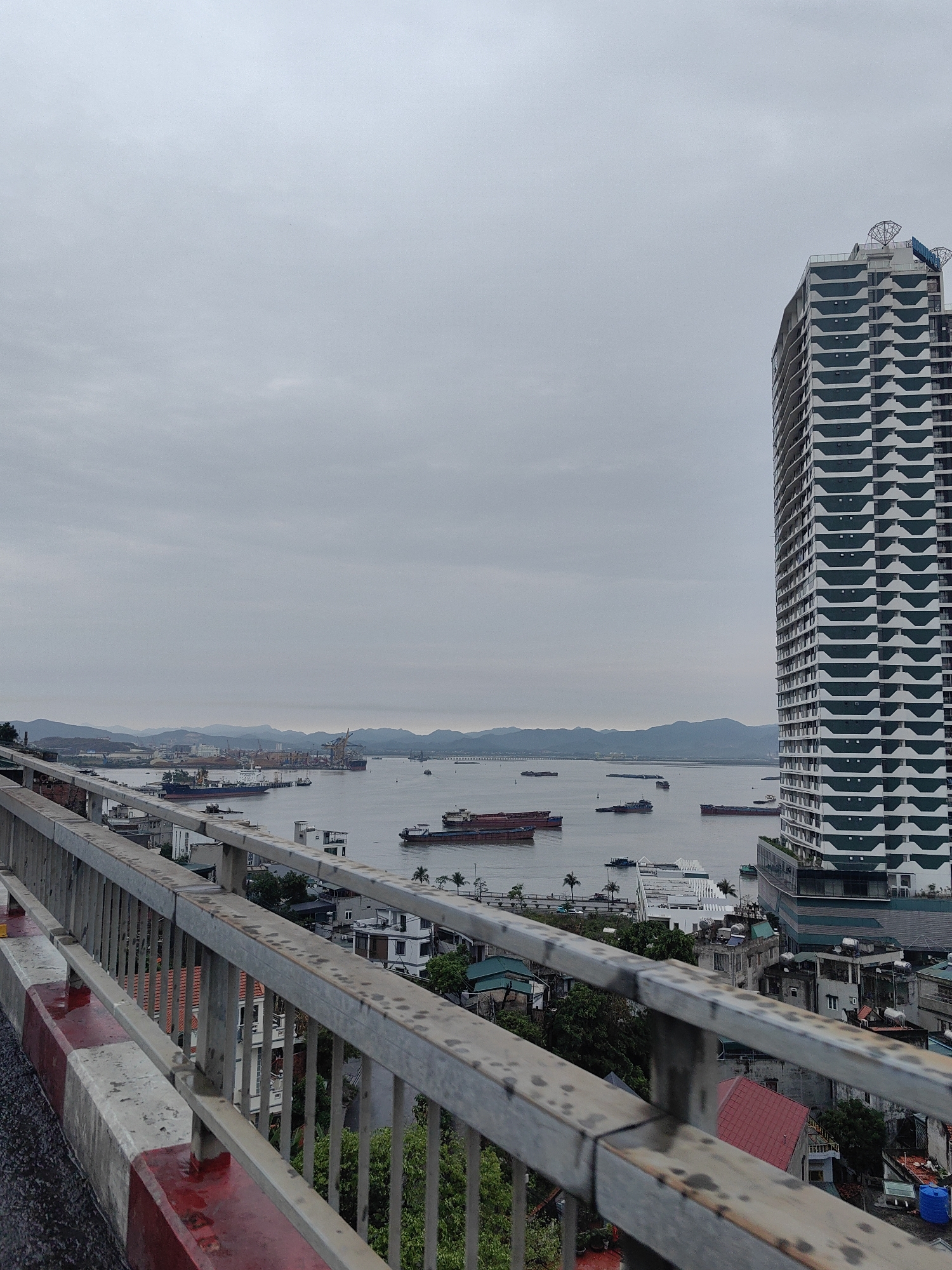 A high-angle view from a bridge overlooking a coastal area in Hong Gai Ward, Vietnam. The foreground features a metal railing painted with alternating red and white stripes on a concrete base. Beyond the railing, a mix of low-rise buildings, trees, and rooftops are visible, extending towards a body of water filled with cargo ships. In the distance, mountains rise against a cloudy sky. A tall, modern building with a white facade and repeating horizontal green accents dominates the right side of the frame. Satellite dishes are visible on top of the skyscraper.