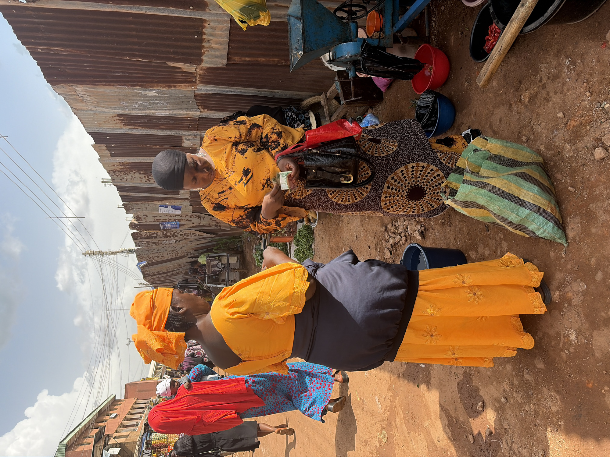 Two women are in a bustling outdoor market in Jos, Nigeria. The scene is characterized by corrugated metal roofing, dirt ground, and a variety of objects commonly found in a marketplace, including plastic buckets, bags, and what appears to be a manual grinding machine.
One woman, wearing a black head covering and a yellow and black patterned dress, is seated. She is holding a banknote in her hand, possibly engaged in a transaction or counting money. In front of her is a black handbag with a gold clasp. The other woman, standing, is dressed in a vibrant orange headscarf and an orange dress with intricate floral embroidery along the hem, layered over a dark grey skirt.
The weather appears to be bright, suggesting it is daytime, likely late morning or early afternoon, given the shadows. The overall impression is one of everyday commerce and activity within a somewhat rustic market environment. The presence of the grinding machine and the general layout suggest a focus on locally produced goods or services. The interaction between the two women, with one holding money, points towards a commercial exchange. The background shows other market stalls and buildings, indicative of a lively and populated area.