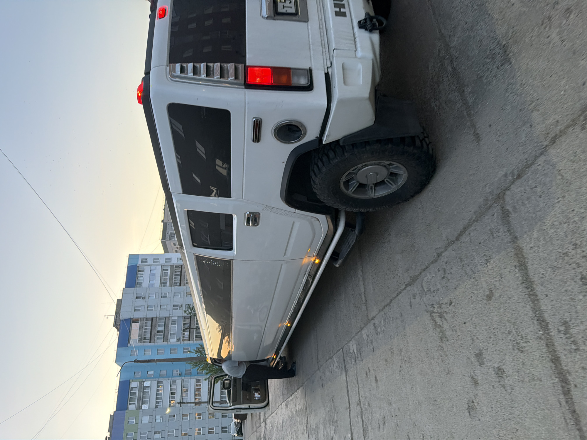 The image shows a white Hummer H2 SUT limousine parked on a concrete street in Chaikovsky, Russia. The vehicle's rear and side are visible, featuring a large tire, a step bar, and open rear door. In the background, a tall residential apartment building with multiple windows and balconies is seen under a slightly hazy sky. Power lines cut across the top left of the frame. A person stands near the open car door. The street surface is cracked and shows signs of wear.