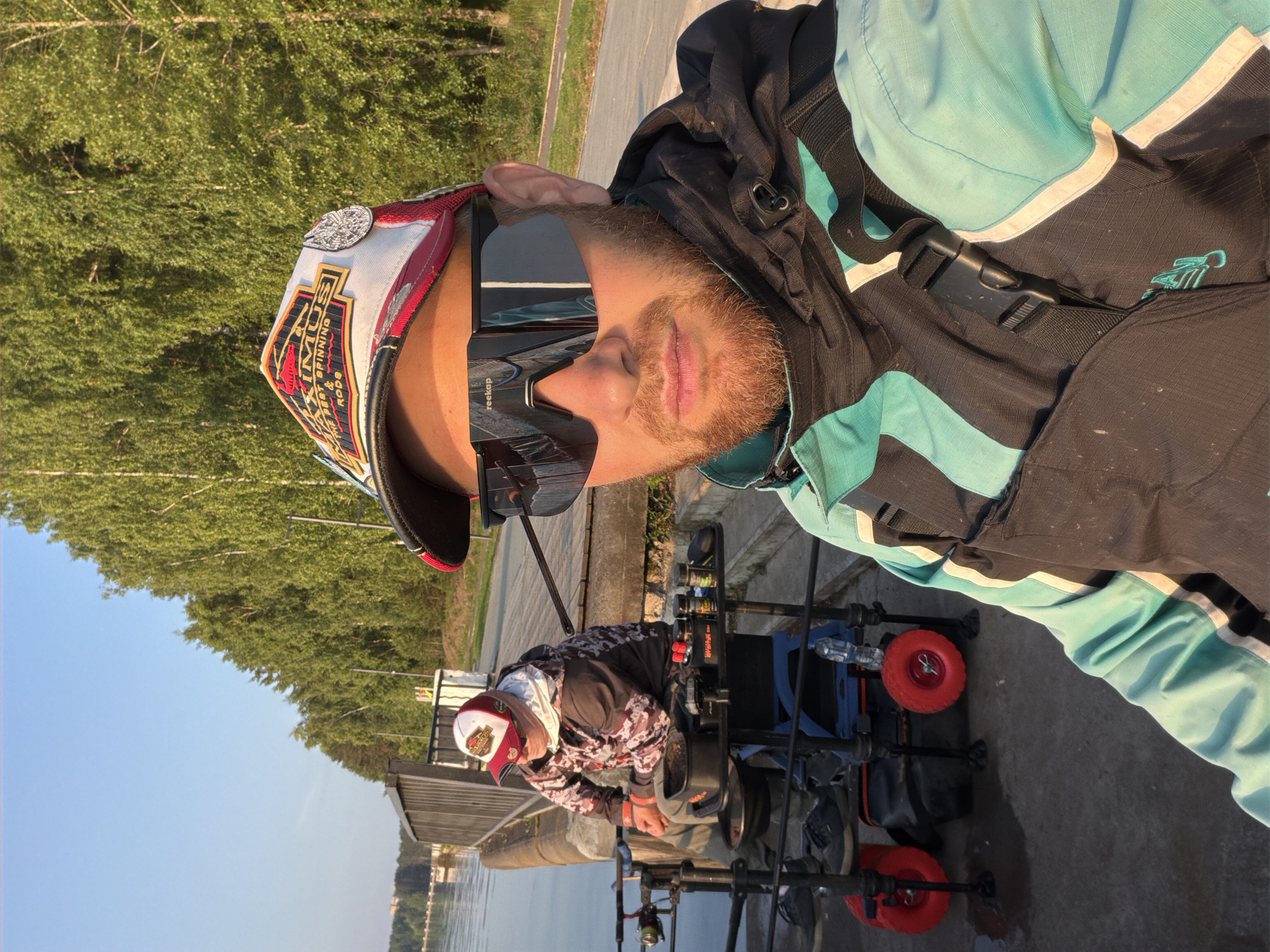 The image depicts two individuals engaged in fishing activities on a pier in Chaikovsky, Russia. The foreground features a man wearing a dark and teal jacket, a cap with multiple patches, and large black sunglasses. He is taking a selfie, holding the camera with his right hand. Behind him, to the left, another person sits on a chair, also wearing a camouflage jacket and a cap. They are surrounded by fishing gear, including fishing rods, a tackle box with various containers, and a portable cart with red wheels. The environment is characterized by a calm body of water, a wooden pier or embankment, and a dense line of green trees in the background, suggesting a natural, outdoor setting. The sun appears to be low in the sky, casting a warm light on the scene.