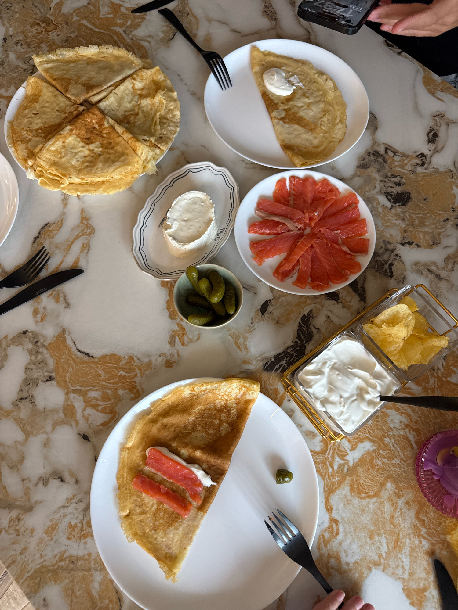 A close-up, top-down view shows a marble table with various food items, suggesting a meal, likely brunch or a light lunch. The setting appears to be indoors, possibly a home dining area in Rostov-on-Don, Russia, given the stylistic elements and presentation.
On the table, several crepes are prominent. One plate holds a stack of crepes, sliced into quarters. Another single crepe is folded on a white plate, topped with a dollop of white cream and two small pieces of smoked salmon. A separate plate displays thinly sliced smoked salmon arranged in a circular pattern. Adjacent to this is a small bowl filled with gherkins. Further to the right, a gold-rimmed container holds sour cream or a similar dip, with a spoon partially submerged in it. Beside the sour cream, another compartment contains potato chips. In the upper right corner, a person's hand is visible, holding a smartphone displaying the time, 17:37. Black cutlery, including forks and knives, are strategically placed around the dishes. A decorative purple item, possibly a flower or dessert, is visible in the far right. The marble surface of the table has a distinct pattern of beige, white, and golden-brown swirls.