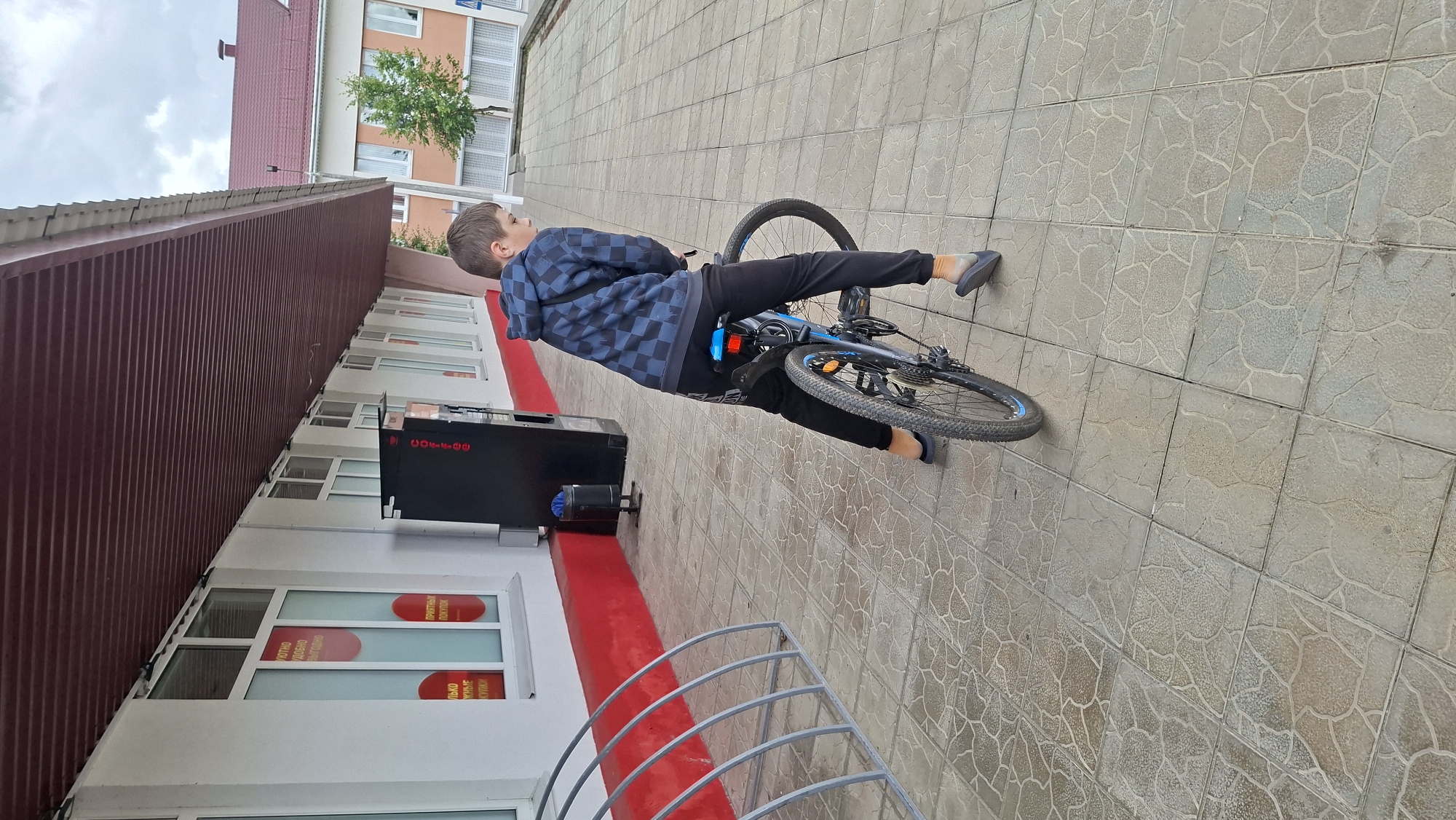 A young boy is standing on a paved sidewalk next to a bicycle. He is wearing a blue and black checkered hoodie and black pants. The bicycle is blue and black. To the left of the boy, there is a bike rack. Behind the boy is a building with white walls and a red trim along the bottom. There are several windows on the building, some of which have red circles with text on them. A black vending machine is also visible next to the building. In the background, there is another building with an orange and white facade and a tree in front of it. The sky is overcast. The image appears to have been taken in Myadzyel, Belarus, given the context of the request.