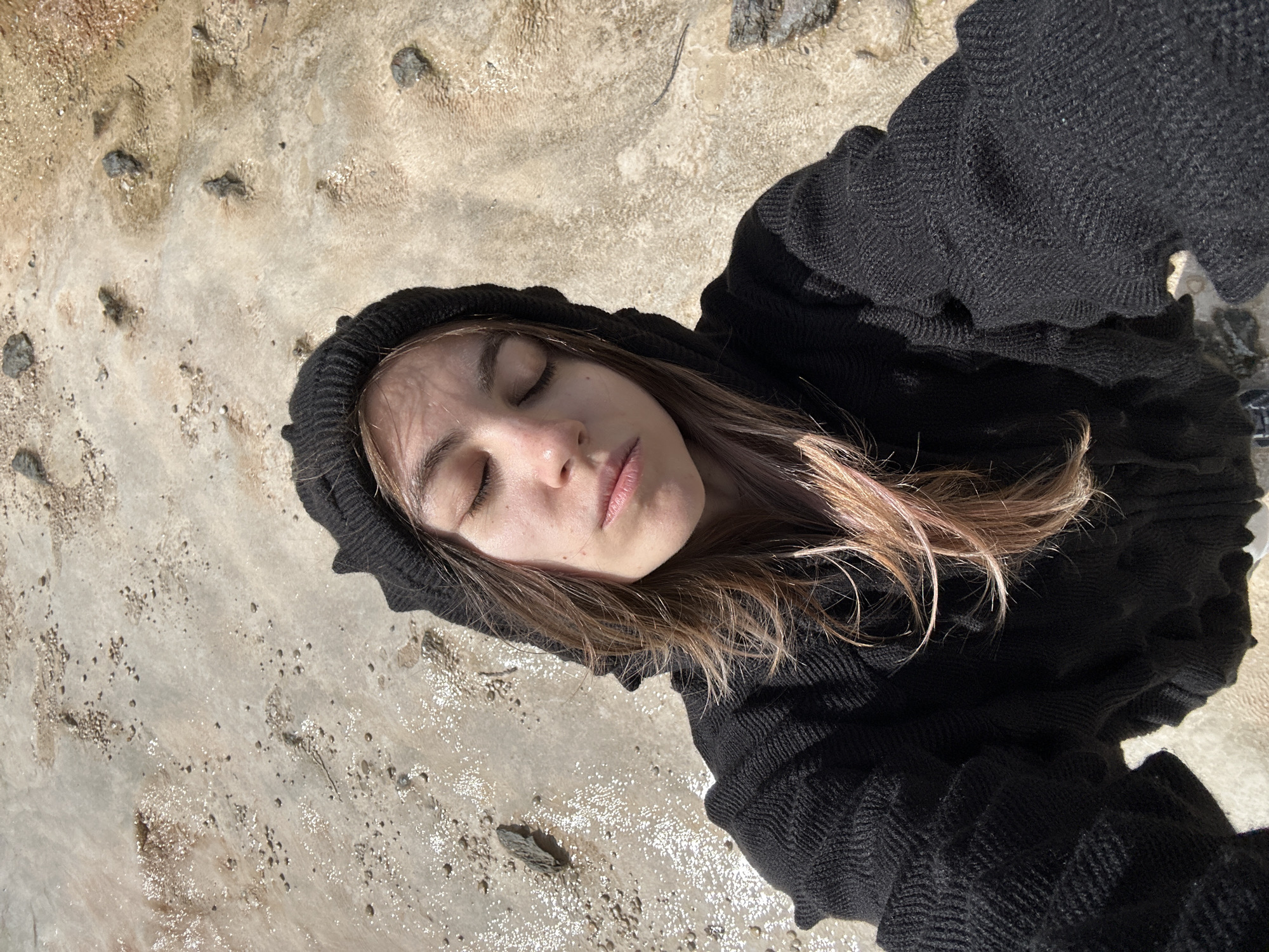 A young woman with her eyes closed and her head tilted back is taking a selfie outdoors. She is wearing a black hooded sweater and has light brown hair with pink highlights. The ground beneath her appears to be a light-colored, textured surface with small pebbles and patches of water reflecting sunlight. The setting does not provide any specific visual cues to identify it as being located in the city of Almasiani, Georgia.
