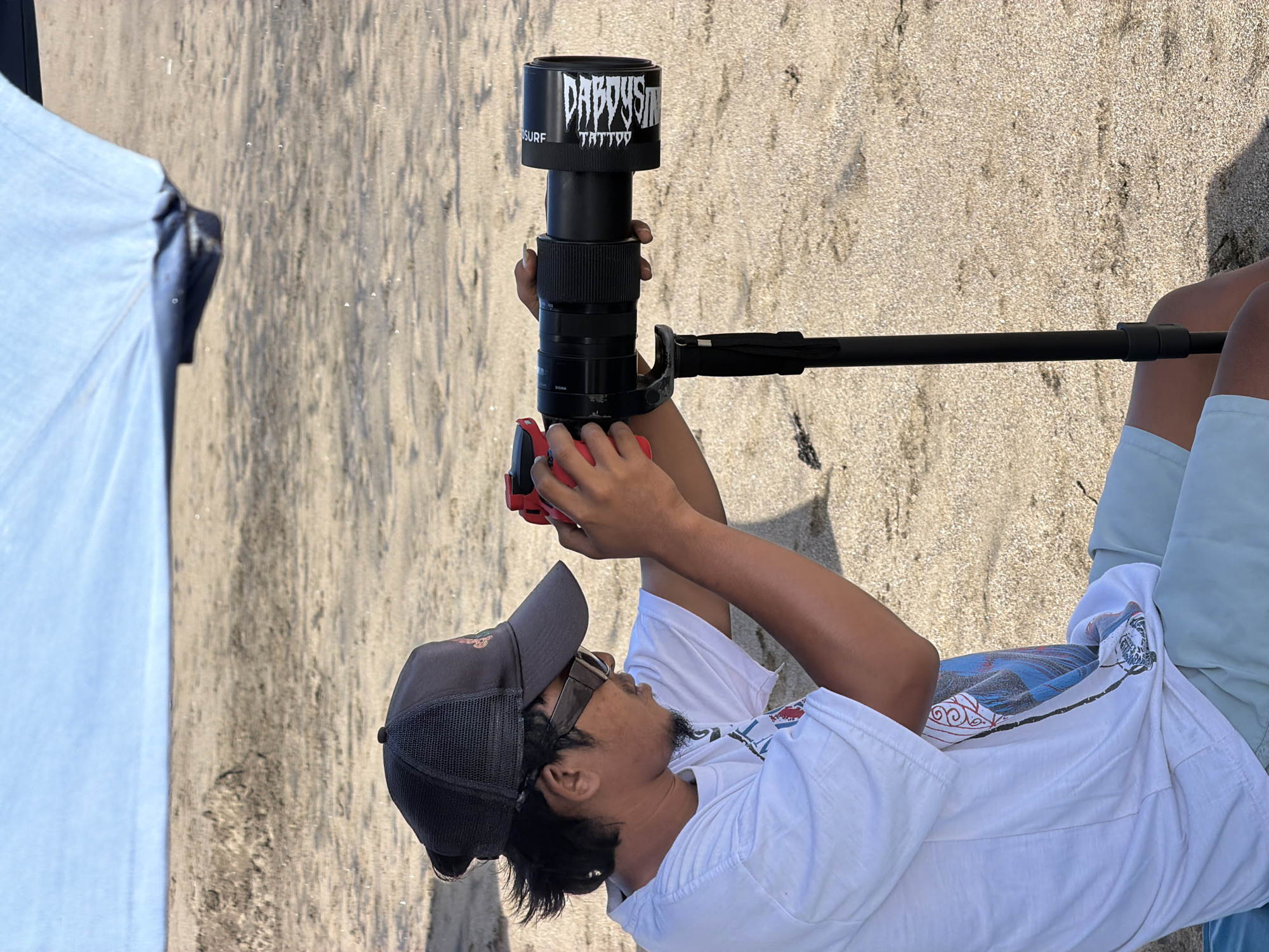 A man wearing a gray trucker hat, sunglasses, a white t-shirt with a graphic print, and light blue shorts sits on a sandy beach under a light blue canopy. He holds a large black telephoto lens with a red camera body attached, which is mounted on a black tripod. The lens has a white sticker with dark lettering that appears to read "DARK VISION TATTOO SURF". The man is looking through the camera, seemingly taking a photograph. The background consists of sand and the blurred ocean. This scene is located on the beach in North Kuta, Indonesia.