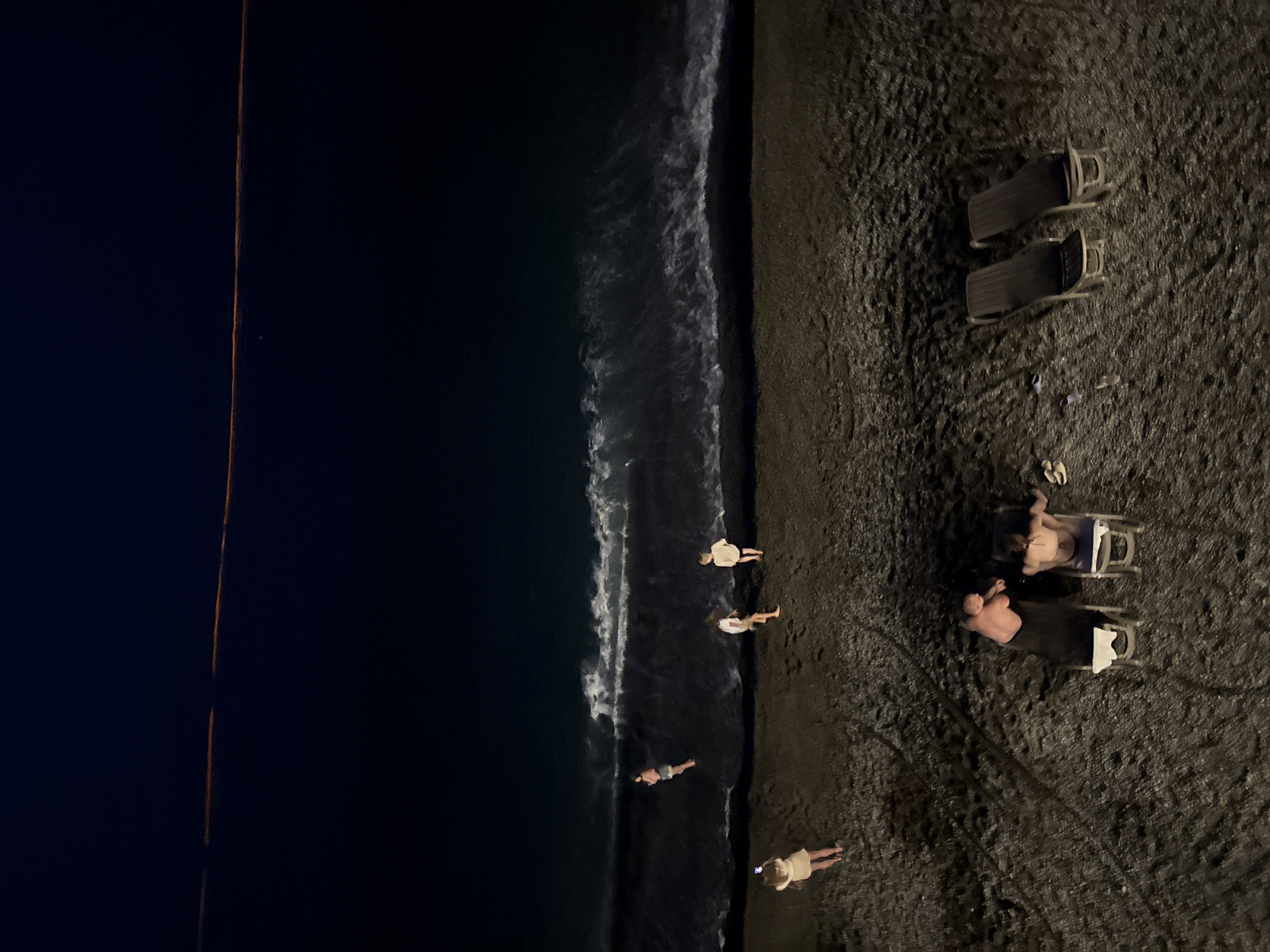 At night on a beach in Yalta, Ukraine, several people are visible. The scene is dark with some lighting, likely from artificial sources, illuminating parts of the sand and water.
In the foreground, two individuals are relaxing on lounge chairs on the dark, pebbly sand. One person, appearing to be an adult male, is lying back on a lounge chair. Another person, also on a lounge chair, is facing away from the camera.
Further up the beach, near the water's edge, four individuals are standing. One person is holding a phone and appears to be taking a picture or video. The other three are facing the ocean. The waves are breaking on the shore, creating white foam against the dark water.
In the distance, a faint orange line is visible across the dark sky, possibly from distant lights or a reflection. The overall atmosphere is tranquil, with the sound of the waves being the most prominent sensory element.