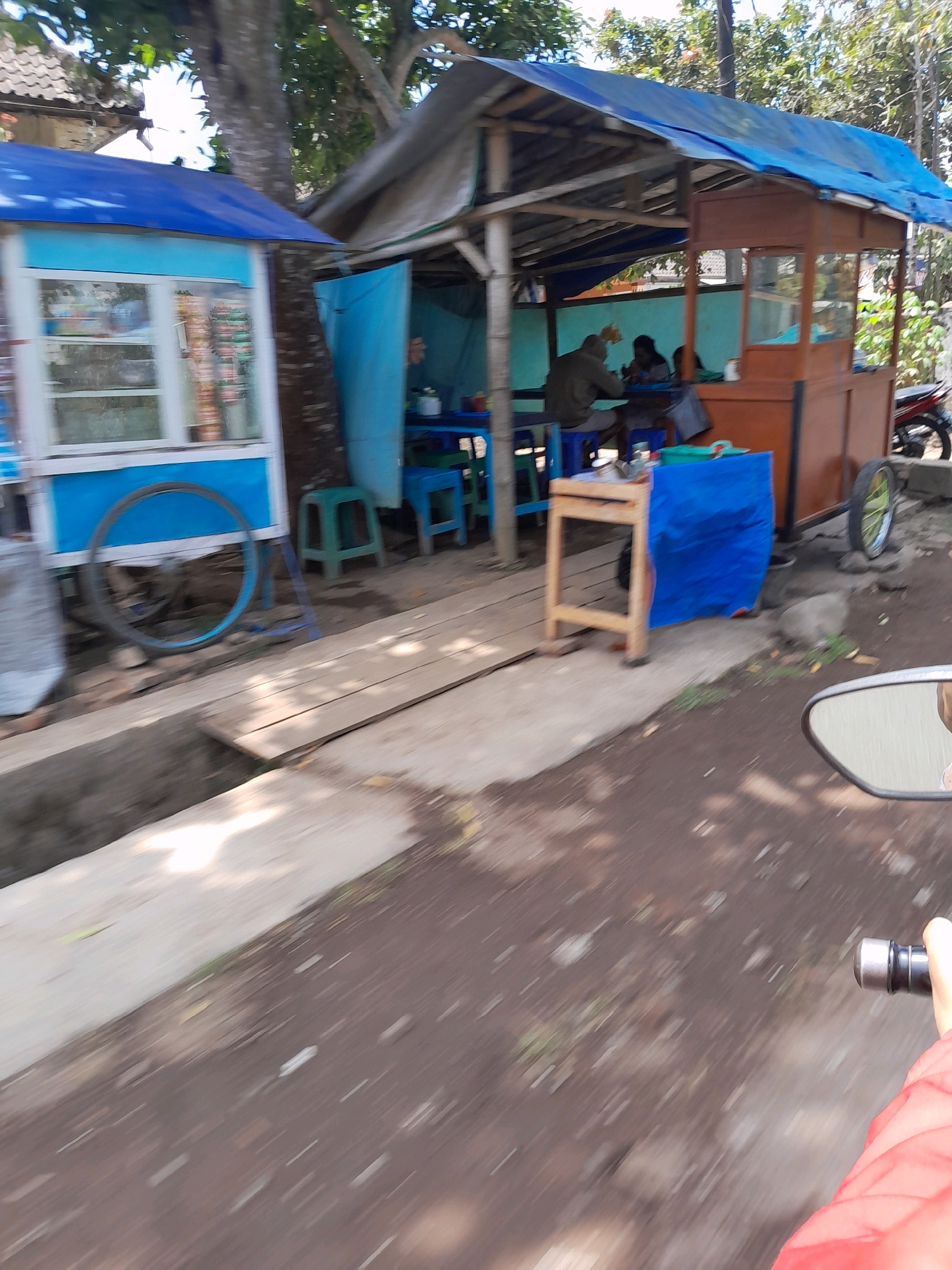 The image depicts a street scene in Jatirejoyoso, Indonesia, featuring two small food stalls or kiosks. The stall on the left is a light blue and white structure with windows displaying various items. To its right, a larger, open-air stall with a blue tarp roof and a wooden counter is occupied by at least two individuals, appearing to be customers or workers. One person is seated at a table, and another is partially visible. A motorcycle is parked nearby. The foreground shows a dirt and concrete ground with some greenery, and the perspective suggests the photo was taken from a moving vehicle, likely a motorcycle, as evidenced by the side mirror in the lower right corner. The overall setting is outdoors, with trees and buildings visible in the background, indicating a somewhat rural or semi-urban environment.