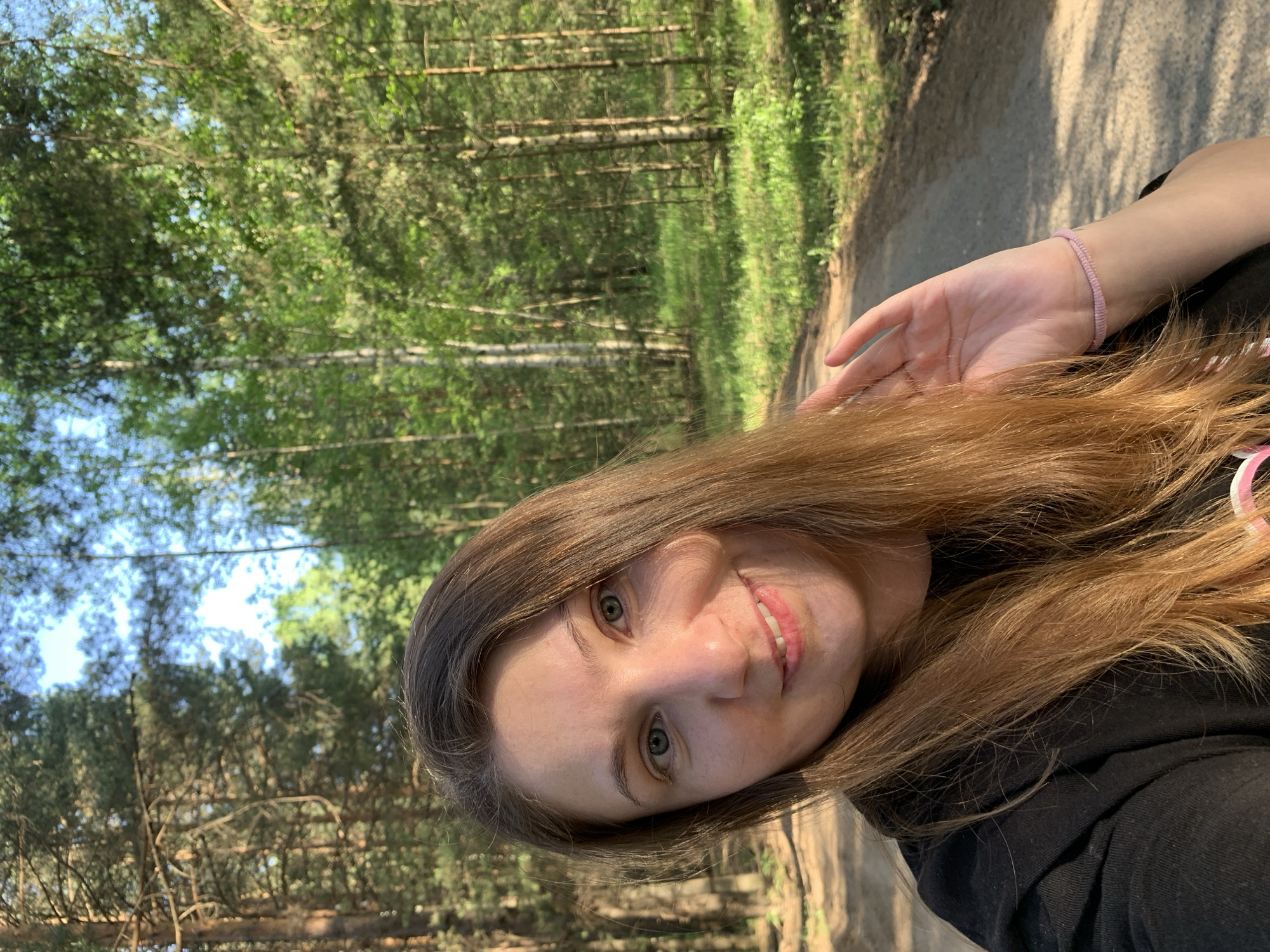 A selfie portrait of a woman with long brown hair, fair skin, and green eyes, positioned on a dirt path within a forest. She is smiling slightly, her hand raised near her face with a light purple bracelet visible on her wrist. She is wearing a black garment. The background consists of dense trees, including birch trees, and foliage, indicating a wooded area. The path appears to be unpaved and well-trodden. The location is assumed to be within the city of Chaikovsky, Russia, based on the prompt.