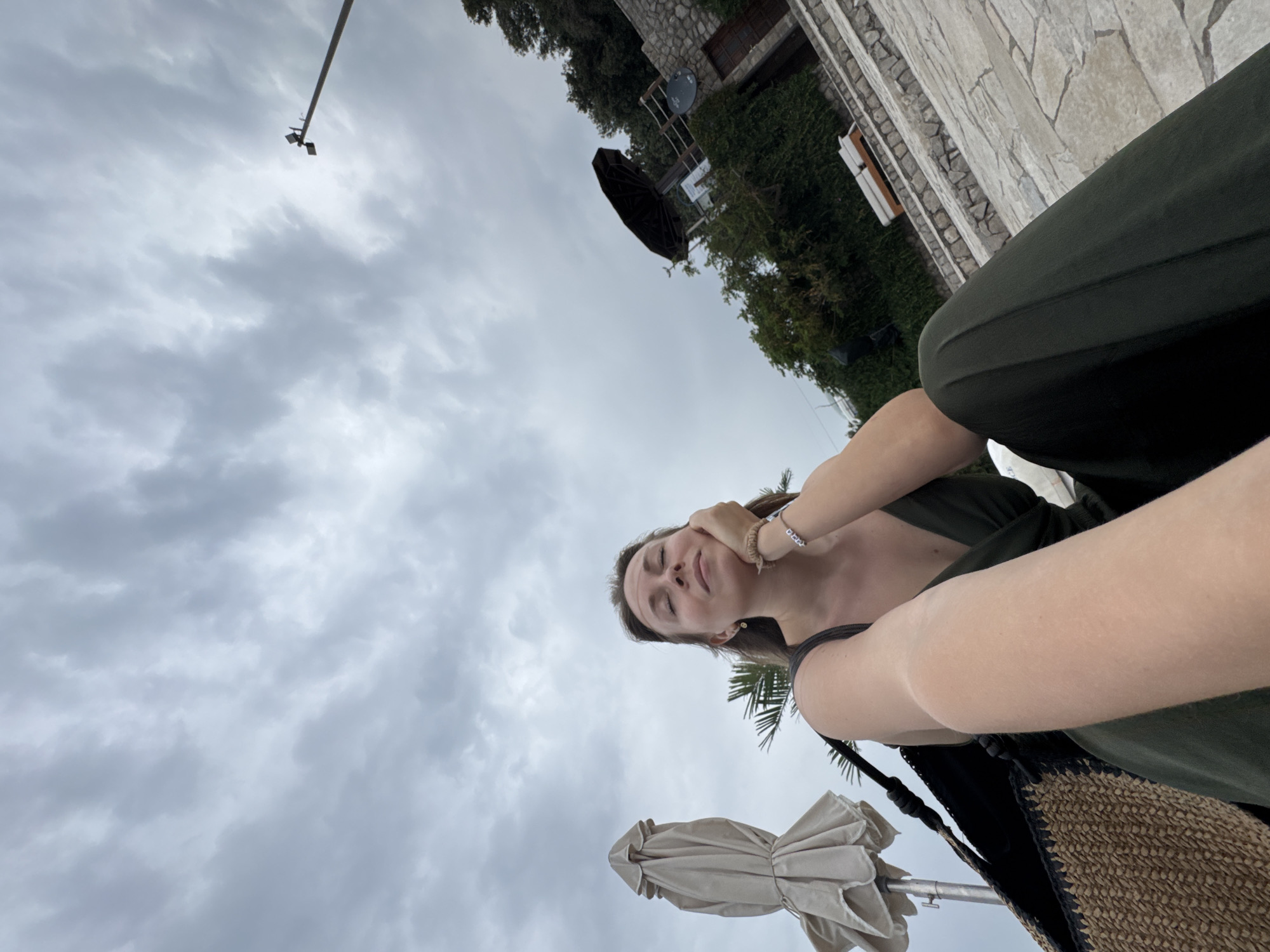 The image, captured from a low angle, features a young woman relaxing outdoors under an overcast sky in Prijevor, Montenegro. The woman, with light brown hair, is shown from the chest up, looking upwards with her eyes closed or cast down in a contemplative expression, and a faint smile. She is wearing a dark green top or dress, complemented by dark green trousers. Her left hand is gently raised to her jawline, revealing a light-colored beaded bracelet on her wrist, with another similar bracelet visible higher on her arm. A small earring is visible in her left ear.
In the foreground, towards the bottom left, a closed beige beach umbrella is visible. Next to the woman, a textured, woven straw or raffia bag with black straps is partially seen. The background consists primarily of a cloudy, grey sky, indicating an overcast day. To the upper right, a stone building with a small window is nestled among dense green trees and foliage. Another large, dark, closed outdoor umbrella is partially visible amidst the greenery. A lamp post or tall pole extends into the sky from the top left of the frame. The scene conveys a sense of calm and relaxation on a cloudy day. There is no visible text in the image.