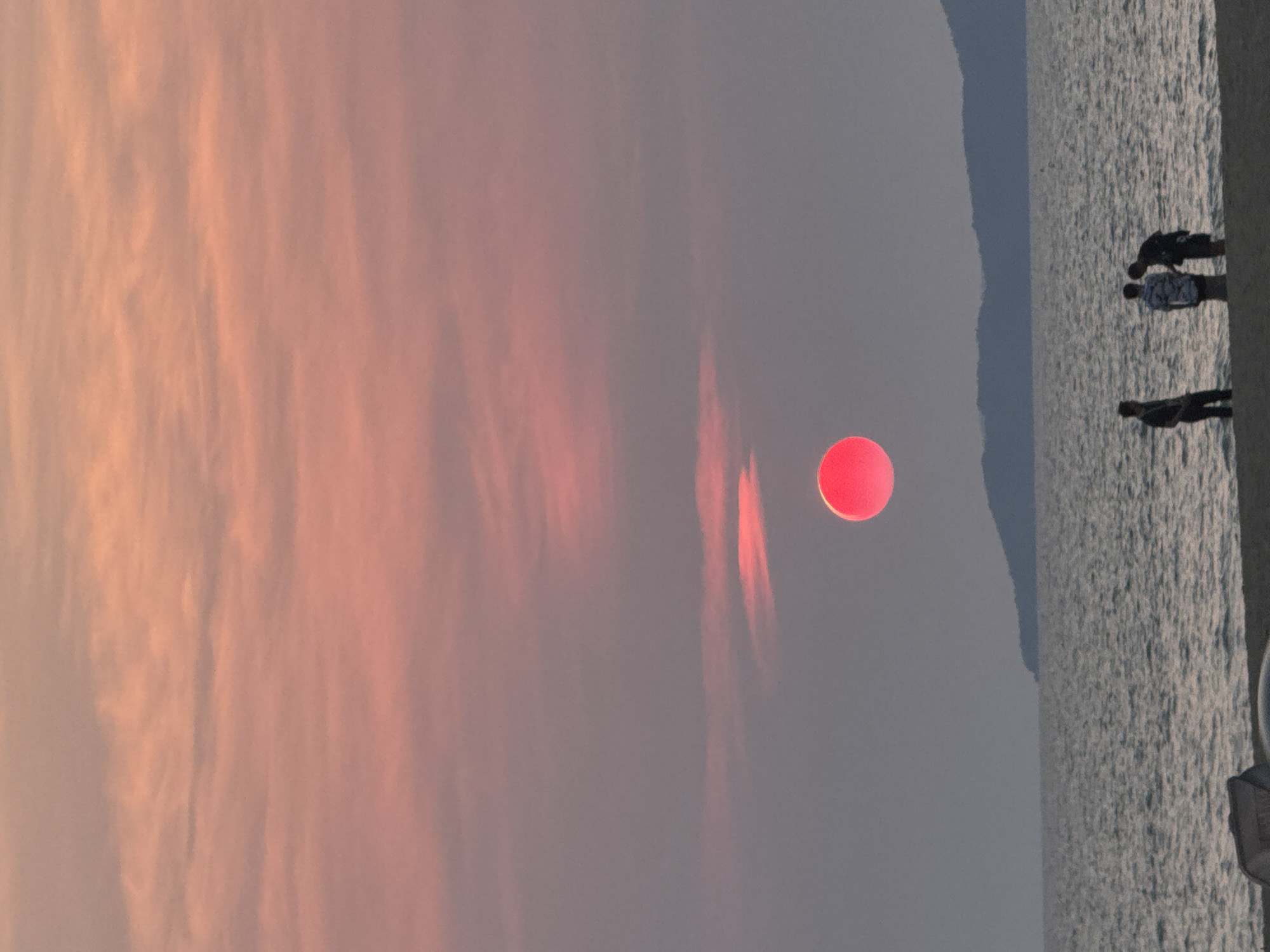 The image captures a sunset scene at a beach in Pattaya City, Thailand. The sky is filled with clouds in the left portion, transitioning to a clear grayish tone. The sun appears as a red sphere, casting light rays. The sea is visible on the right, and a vague silhouette of a coastal mountain range. Several people stand at the edge of the water on the beach.