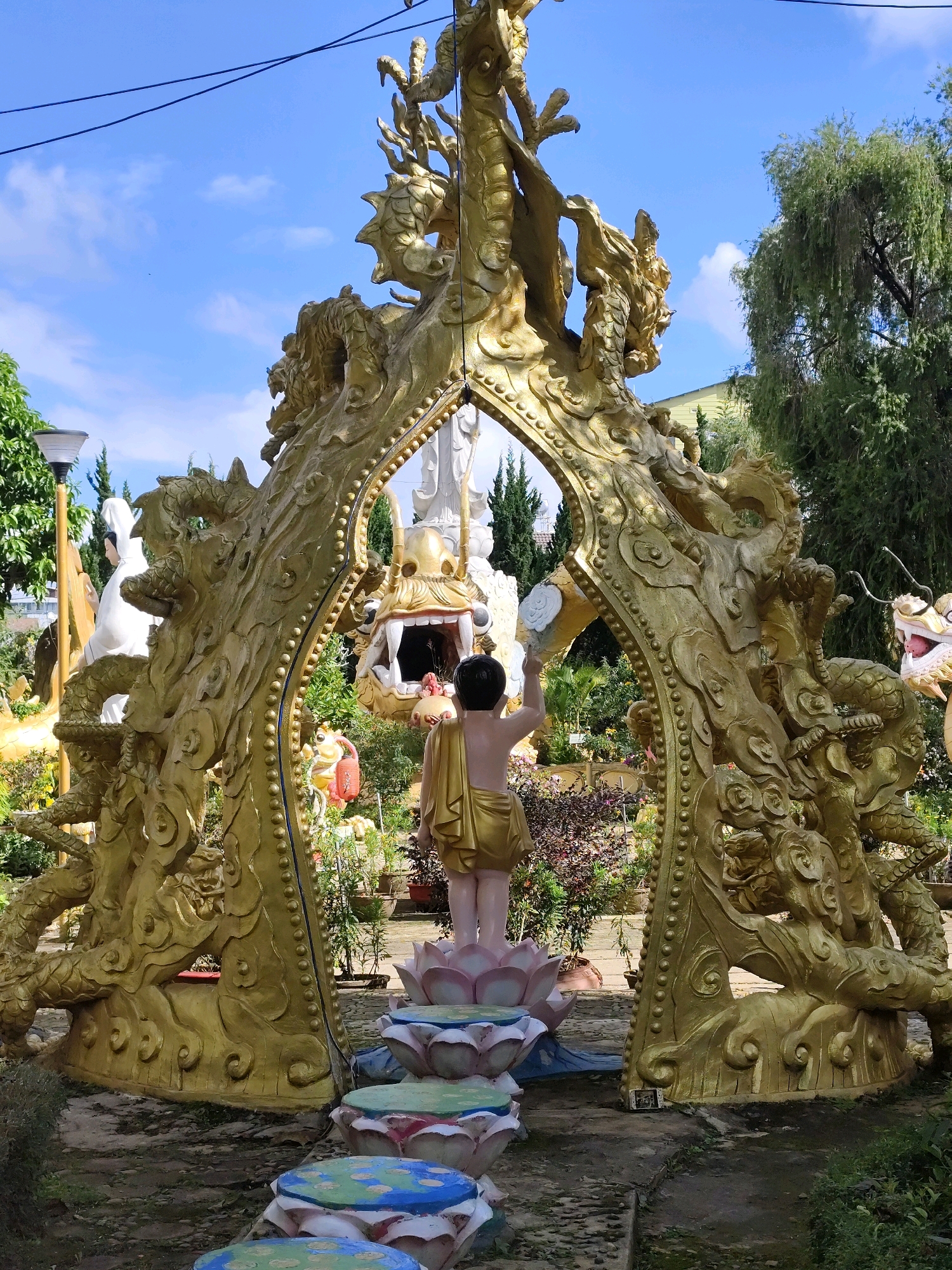 This is a medium shot, eye-level, outdoor, full-color photograph captured in the city of Da Lat, Vietnam, possibly at the Thien Vuong Pagoda or a similar Buddhist temple complex. The central focus is a large, golden, ornate archway adorned with sculpted dragons. The archway has a distressed, weathered appearance, suggesting it is aged. Within the arch, a white statue of a serene figure, possibly Buddha or a Bodhisattva, is visible, partially obscured by the golden dragon sculptures. In front of the archway, a smaller statue of a child wearing a golden robe stands, holding a white object, possibly a cup or jug, and appearing to pour a liquid. The child is positioned on a lotus flower base, with a series of layered, colorful lotus-shaped steps leading away from it, painted with blue, green, and pink accents. The background reveals lush greenery, including trees with drooping branches, and other statues and decorative elements characteristic of a temple garden. A bright blue sky with scattered white clouds forms the backdrop. The ground is covered in gravel and dirt, with some patches of grass. A discarded white mobile phone rests on the ground near the base of the archway, to the right of the frame. The overall atmosphere suggests a religious or spiritual site with a strong emphasis on Buddhist iconography.