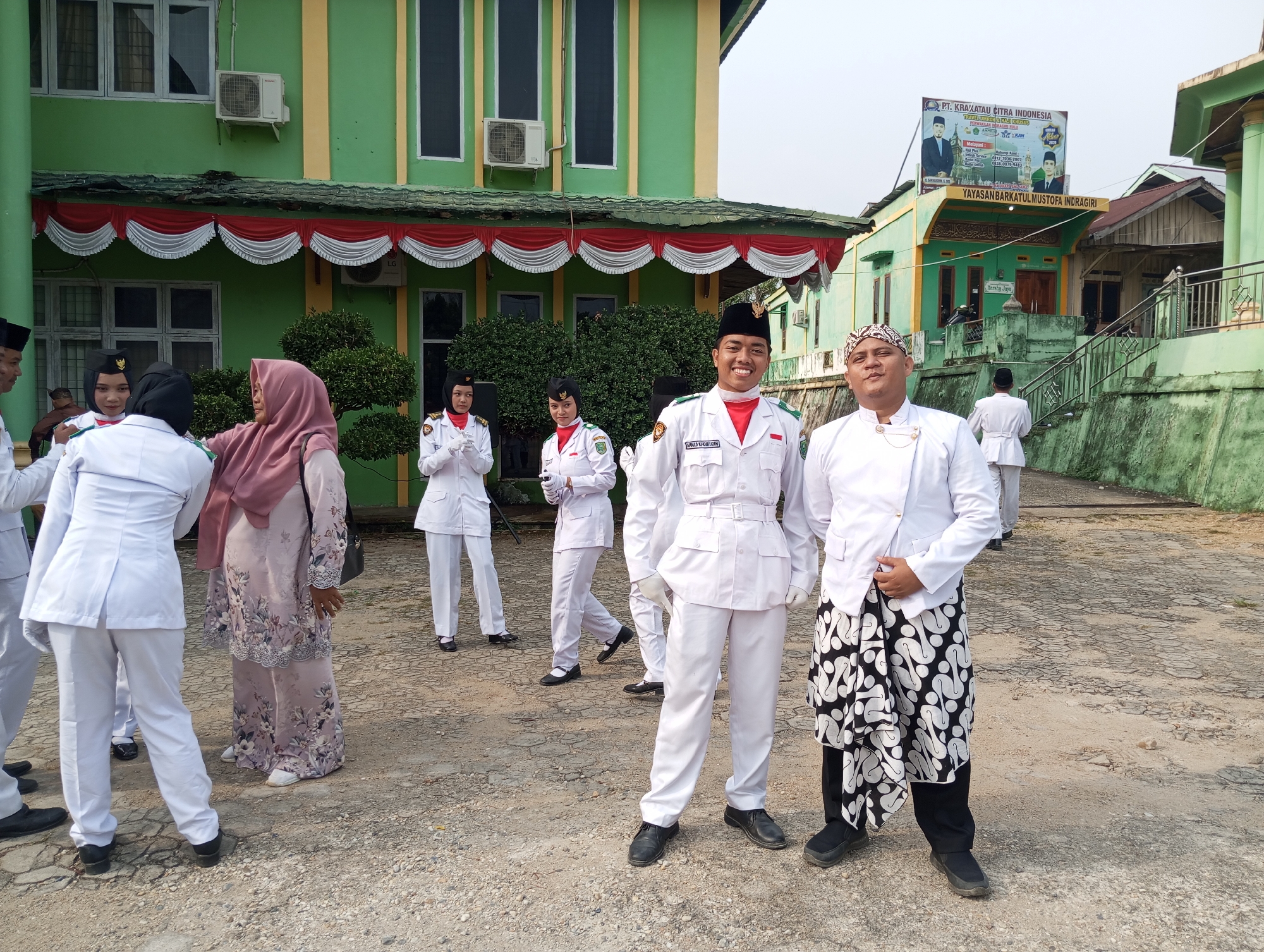 The image depicts an outdoor scene in front of a two-story green building, likely a school or administrative office, in Pematang Reba, Indonesia. Several individuals, predominantly in white uniforms, are visible. In the foreground, two men in white uniforms and traditional headwear are posing for the camera. One of the men is smiling broadly and wearing a black cap with an emblem. The other man is wearing a patterned headscarf and a white jacket over black pants with a white batik sarong.
Behind them, a group of people in white uniforms are gathered, some wearing black caps and others wearing hijabs. One woman in a floral pink hijab stands slightly apart, facing away from the camera. The building's facade features red and white decorative bunting along the roofline. Air conditioning units are mounted on the exterior wall. The ground is paved with gravel and cracked asphalt. In the background, a billboard with Indonesian text and images of men is affixed to a yellow structure above the building. Other buildings and trees are also visible in the background. The sky is overcast.