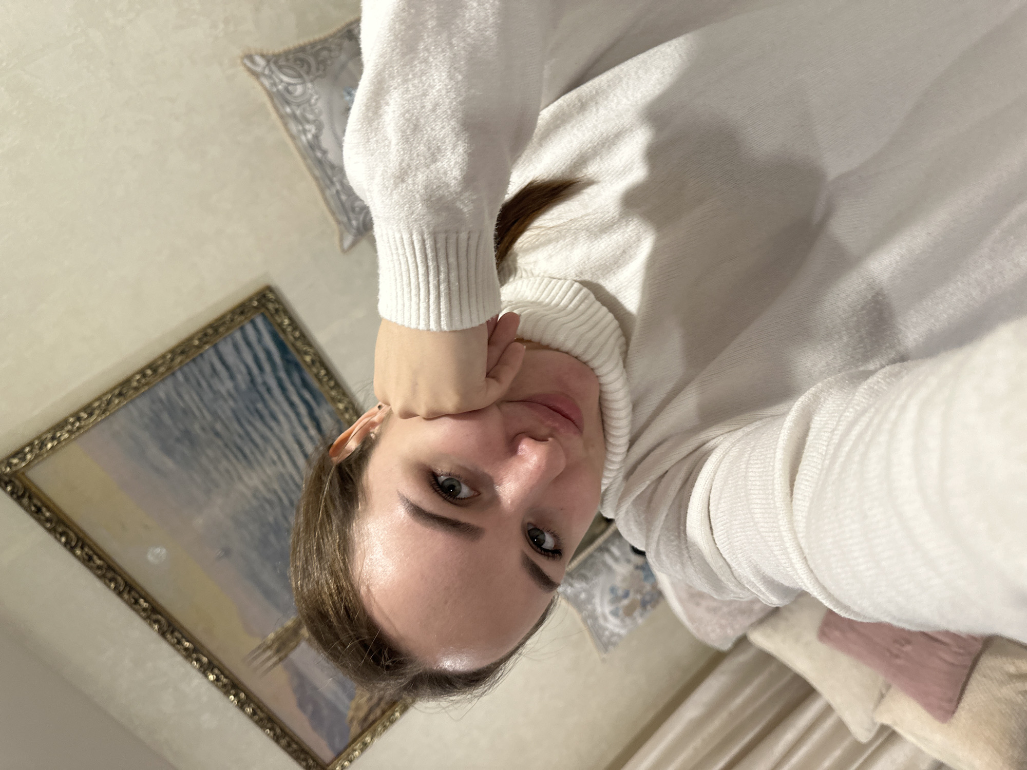A selfie-style portrait of a fair-skinned woman with light brown hair is the central subject. She is wearing a white turtleneck sweater and her face is positioned in the lower-center of the frame. Her left hand is raised to her face, with her chin resting on her fist.
Behind her, on the wall, is a framed landscape painting with a blue and white color scheme and an ornate gold frame. To the upper-left of the woman's head is a decorative pillow with a grayish-blue pattern.
In the background, partially visible, are additional pillows in light colors, including beige and pink. Drapes are seen along the bottom edge of the image.
The location appears to be indoors, possibly a bedroom or living room. There is no verifiable evidence to confirm that the location is in the city of Chaikovsky, Russia.
