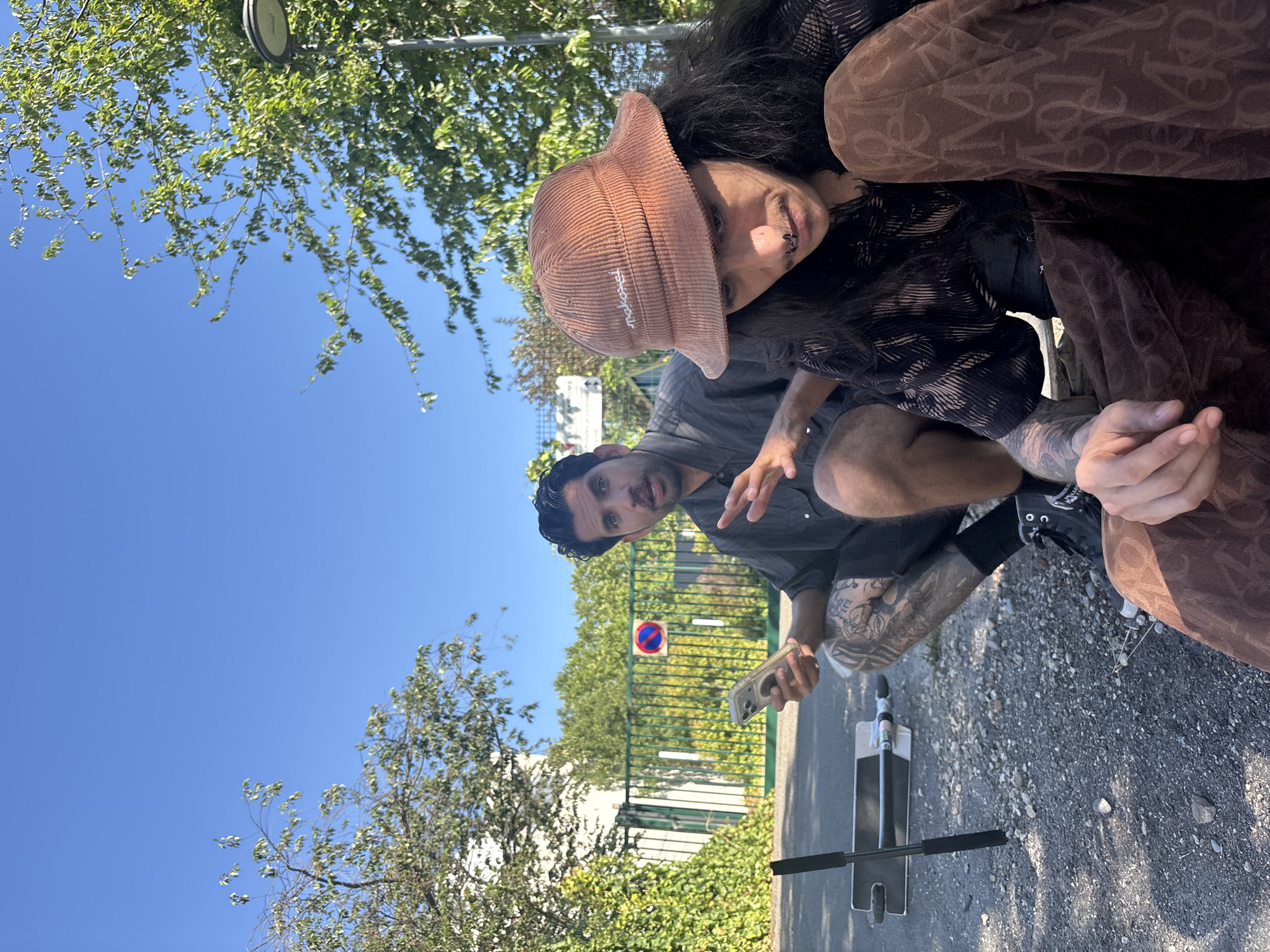 In an outdoor scene under a clear blue sky, two men are crouched on a gravel surface. The man on the left, with dark hair and a beard, wears a dark short-sleeved button-down shirt and is holding a smartphone. His right arm is raised and gesturing. He has extensive tattoos on his legs. In the background, a green metal fence is visible, with a building behind it and lush green trees. A circular sign with a red border and a blue center indicating a prohibition is attached to the fence. A scooter is positioned on the left side of the frame. The man on the right, with long dark hair and wearing a brown corduroy bucket hat, is looking down and to his right. He is wearing a dark patterned shirt and brown pants with a repeating pattern of letters. His nose is pierced. The scene suggests an urban or suburban outdoor setting, possibly in Lyon, France, given the context provided.