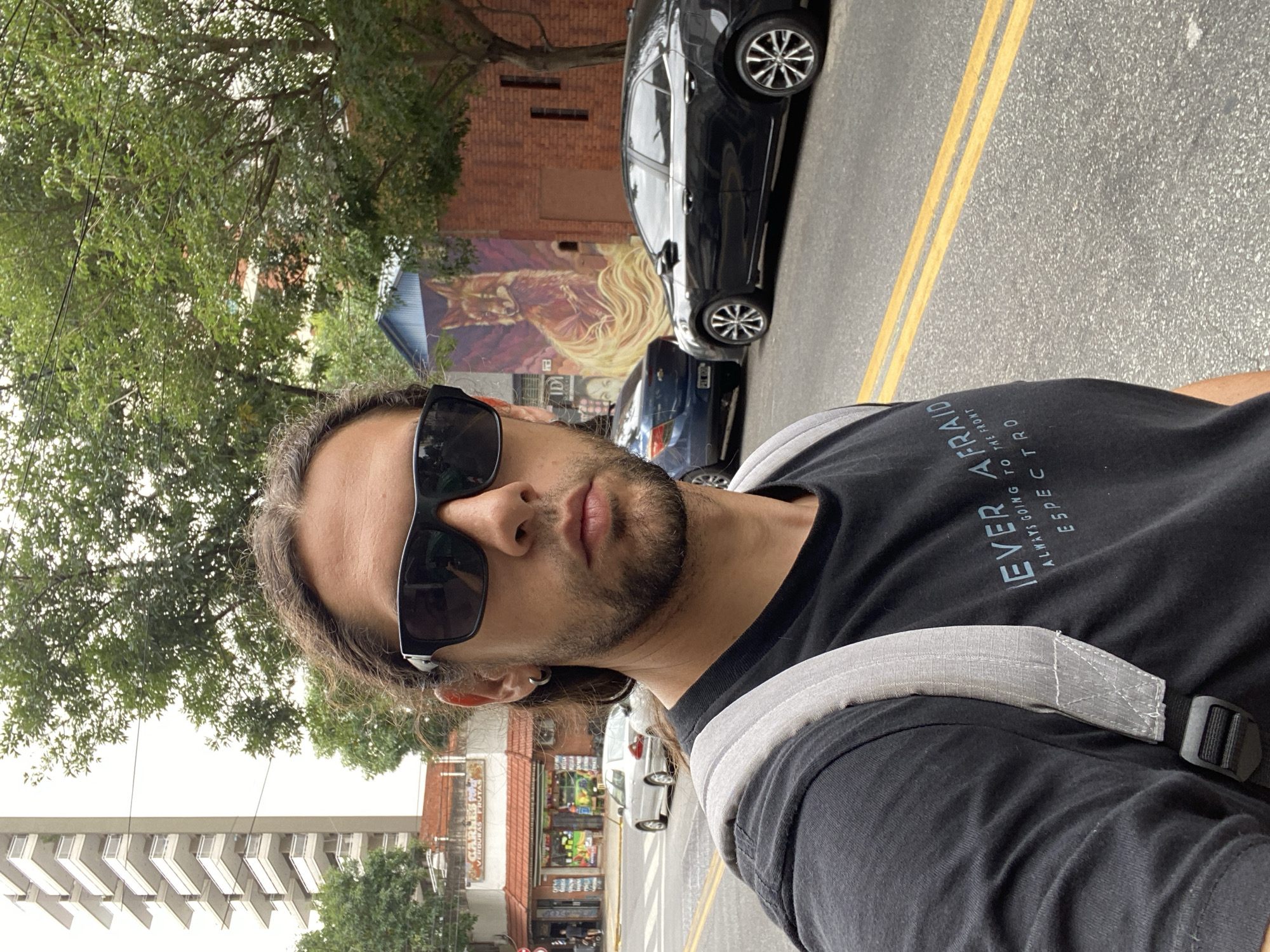 The image depicts a male individual taking a selfie on an urban street in the city of Buenos Aires, Argentina. The subject occupies the left half of the frame, facing the camera directly. He has shoulder-length dark hair, a beard, and wears black rectangular sunglasses and a dark grey t-shirt with partial white text visible: "NEVER AFRAID," "ALWAYS GOING TO THE FRONT," and "ESPECTRO." A light grey backpack strap is visible over his right shoulder.
The street, paved with asphalt, features double yellow lines. In the background to the left, a multi-story, light-colored apartment building with numerous balconies rises above a smaller building at a street corner. This corner building houses a shop identified by the sign "CARLOS VERDURAS Y FRUTAS" (Carlos Vegetables and Fruits) and has an orange-tiled roof. A silver sedan is visible on the street near this corner, and a pedestrian is partially visible on the sidewalk.
To the right of the subject, a large green-leafed tree partially obscures the background. Beneath the tree, a building with a reddish-brown brick facade displays a vibrant mural of a fox-like creature with a long, flowing golden-orange tail. This building features a blue-colored roof section. Two parked sedans are visible on the street to the right: a dark blue vehicle followed by a black sedan with distinct spoked wheels. The scene is illuminated by daylight under an overcast sky.