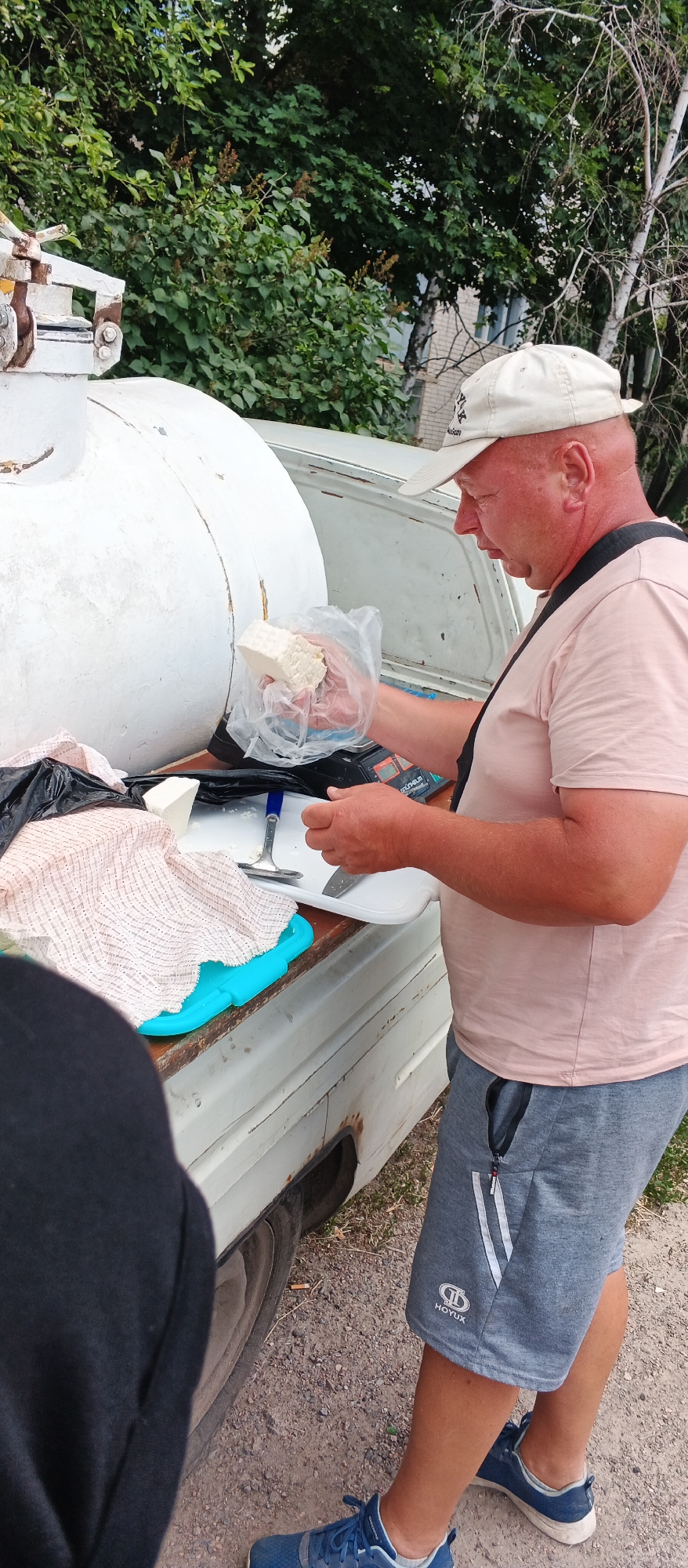 In the city of Smila, Ukraine, a middle-aged man wearing a light-colored baseball cap, a pink t-shirt, and grey shorts stands by the open back of a white truck. He is holding a block of white cheese in a plastic bag in his right hand, reaching to place it on a white plate positioned on a wooden surface. A digital scale is visible beneath the plate. Next to the plate are a blue plastic mat and a patterned cloth draped over a black bag. To the left, a person wearing a dark hoodie is partially visible. The background consists of green trees and vegetation.
