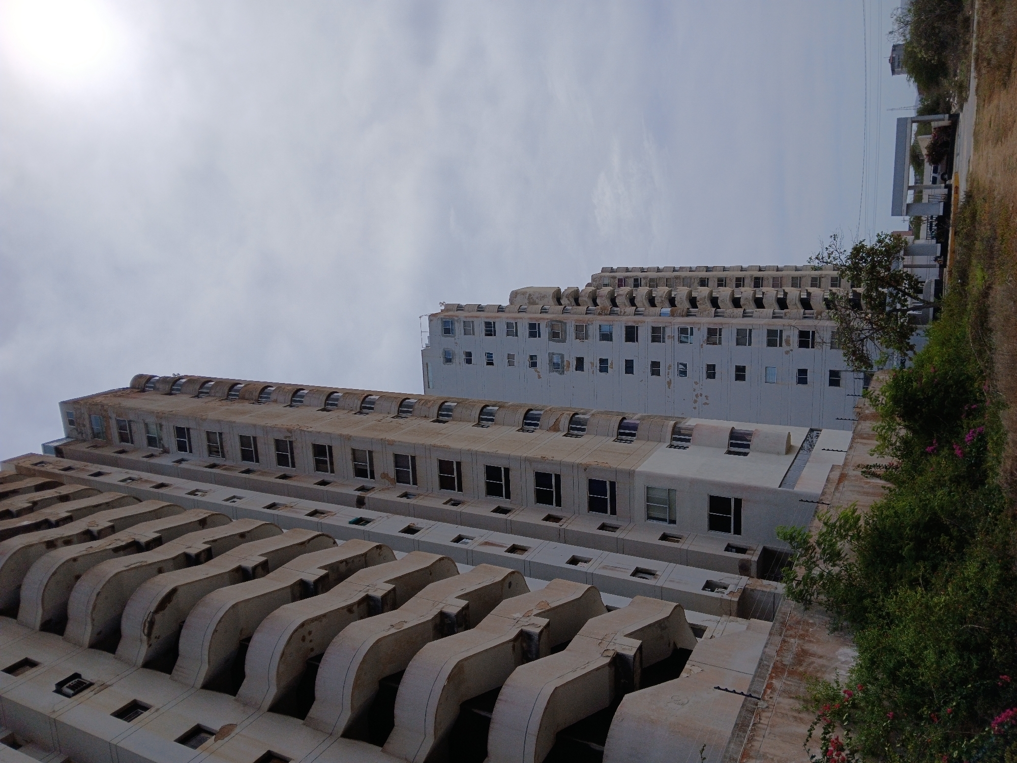 The image shows two tall, aged buildings in Porlamar, Venezuela, viewed from a low angle. The building on the left has distinct wavy, concrete architectural elements protruding from each floor. The building behind it is simpler in design, with rows of windows. The buildings are weathered and show signs of disrepair. To the right of the buildings, there's some greenery and partial views of other structures in the distance. The sky is overcast.