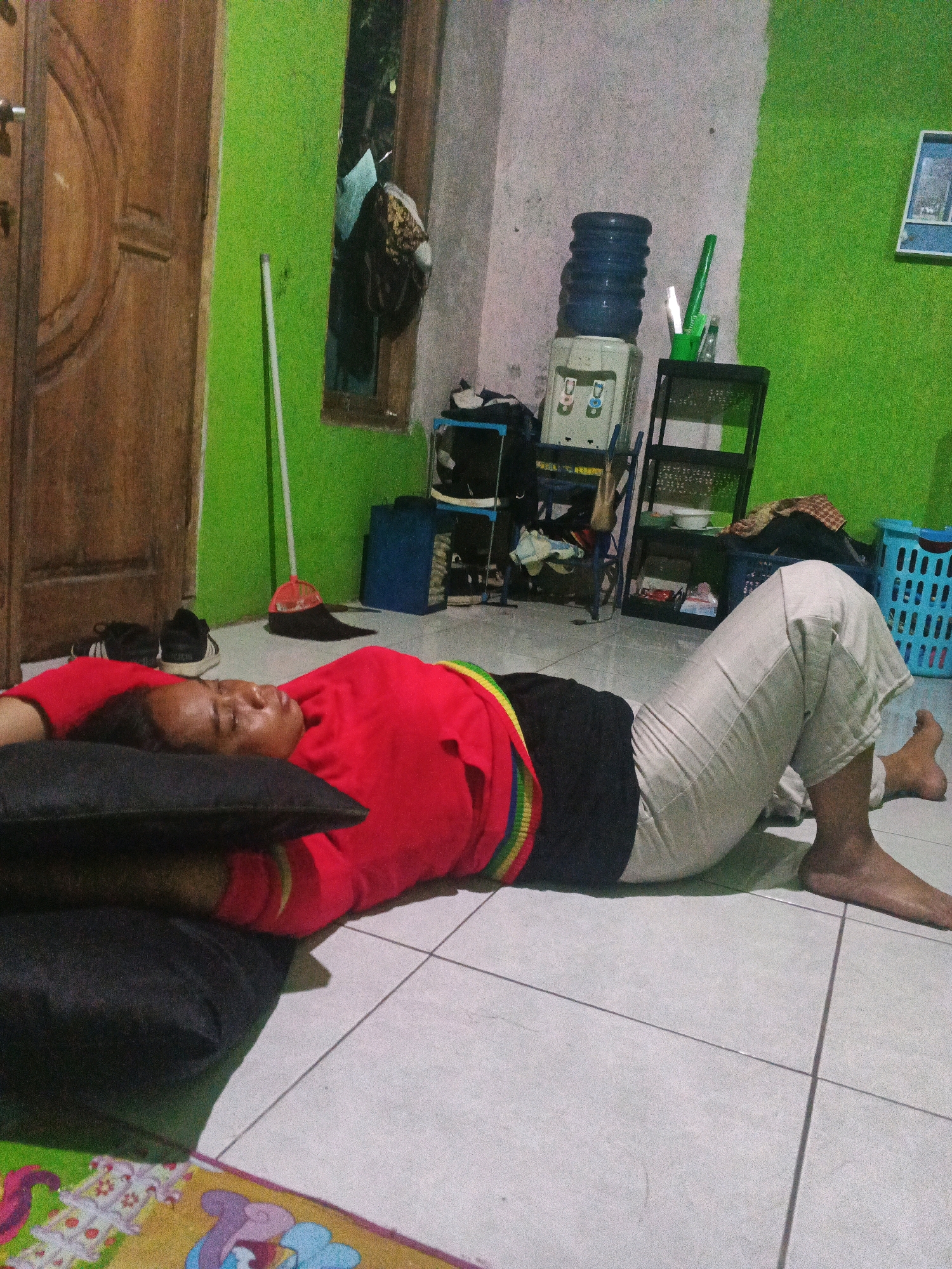 A person lies on the floor on their back with their head resting on a dark pillow. They are wearing a red long-sleeved shirt and light-colored pants. The person's eyes are closed. The floor is tiled. To the left of the person is a wooden door and a green wall. To the right, there is a water dispenser, shelves with various items, and a blue laundry basket. The scene appears to be indoors, likely in a residential setting. This image is believed to have been taken in the city of Semarang, Indonesia.