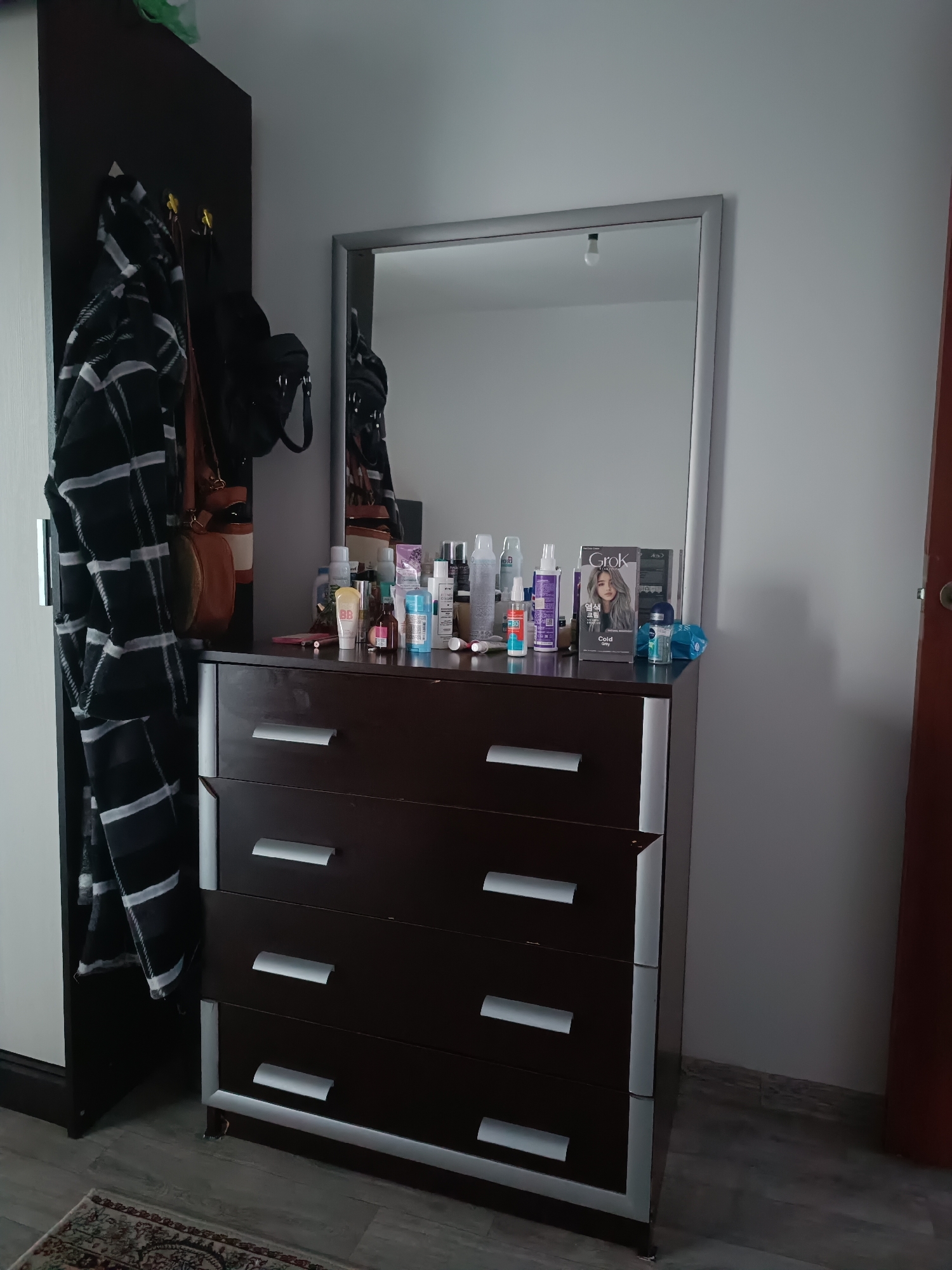 The image displays a dresser with a mirror above it, set against a neutral gray wall. The dresser is dark brown with silver trim and has three drawers, each with a horizontal silver pull. On top of the dresser, there are various toiletries and cosmetics, including bottles, tubes, and a box of hair dye. To the left of the dresser, a dark wardrobe is partially visible, with a black and white patterned garment hanging on hooks. A brown handbag and another dark bag are also hanging on the hooks. A glimpse of a colorful rug can be seen on the floor in the lower left corner of the image. The scene appears to be in a domestic setting, likely a bedroom or dressing area.