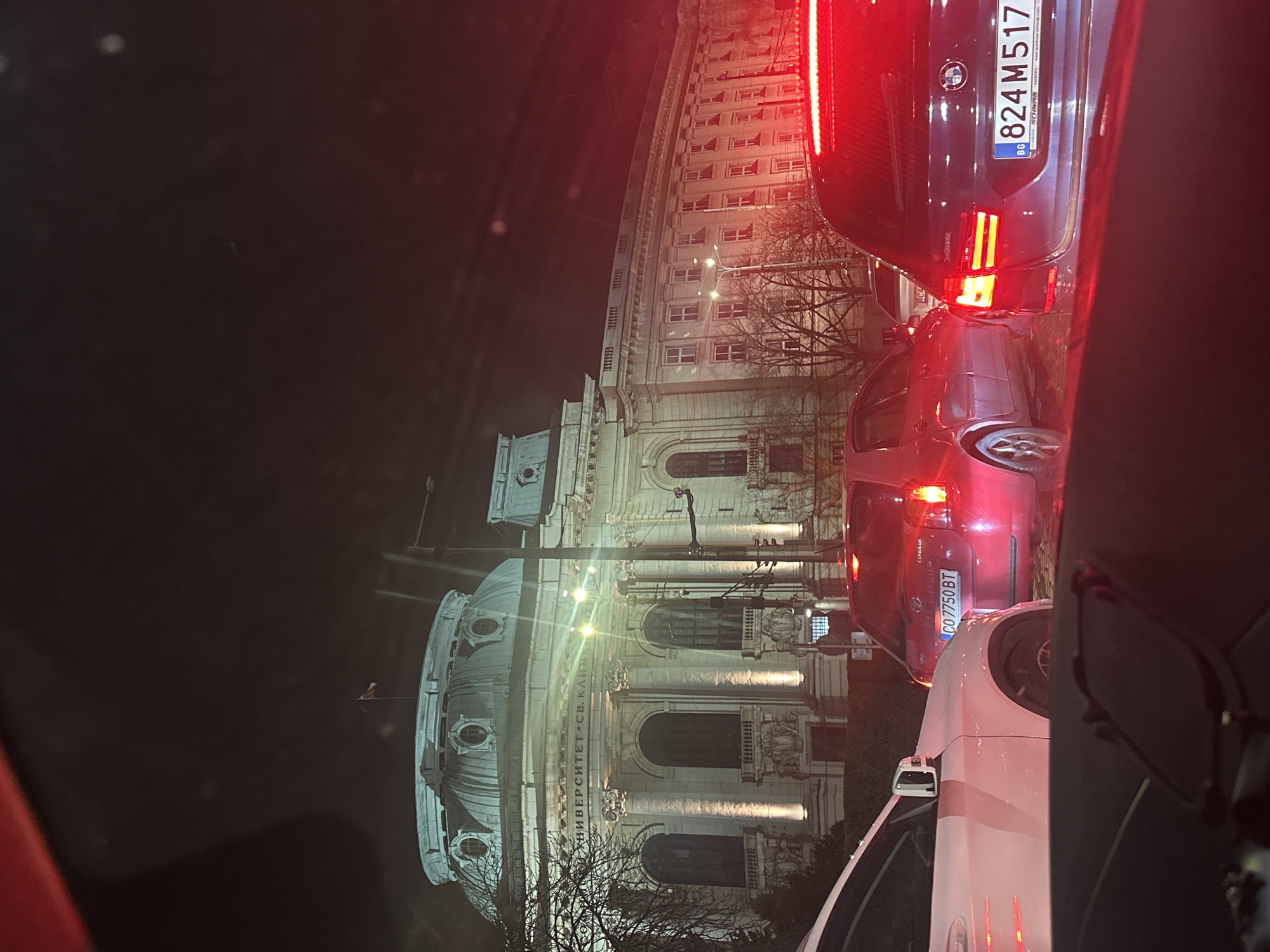 The image captures a nighttime urban scene in Sofia, Bulgaria, depicting stationary vehicular traffic with the illuminated Sofia University "St. Kliment Ohridski" in the background.
The perspective is from inside a vehicle, looking forward through a windshield, with a dark dashboard and red reflections visible in the foreground. Three vehicles are prominently featured: on the far left, the front quarter panel of a white vehicle is partially visible. In the midground, a red Toyota Corolla hatchback (Bulgarian license plate CO 7750 BT) is stopped with its brake lights illuminated. To its right, a dark blue/grey BMW station wagon (Bulgarian license plate BG 824 M517) is also stopped with its brake lights active.
The background is dominated by the floodlit, classical facade of Sofia University "St. Kliment Ohridski," identifiable by the inscription "УНИВЕРСИТЕТ "СВ. КЛИМЕНТ ОХРИДСКИ"" (University "St. Kliment Ohridski") above its central entrance. The building features a large central dome with circular windows and a Bulgarian flag flying from a flagpole on its apex. Columns with ornate capitals and arched windows characterize the main section. To the right, a multi-story wing of the university building extends, featuring numerous rectangular windows. Bare deciduous trees line the street in front of the university. The sky is dark, indicating night, with various streetlights and vehicle lights contributing to the scene's illumination.