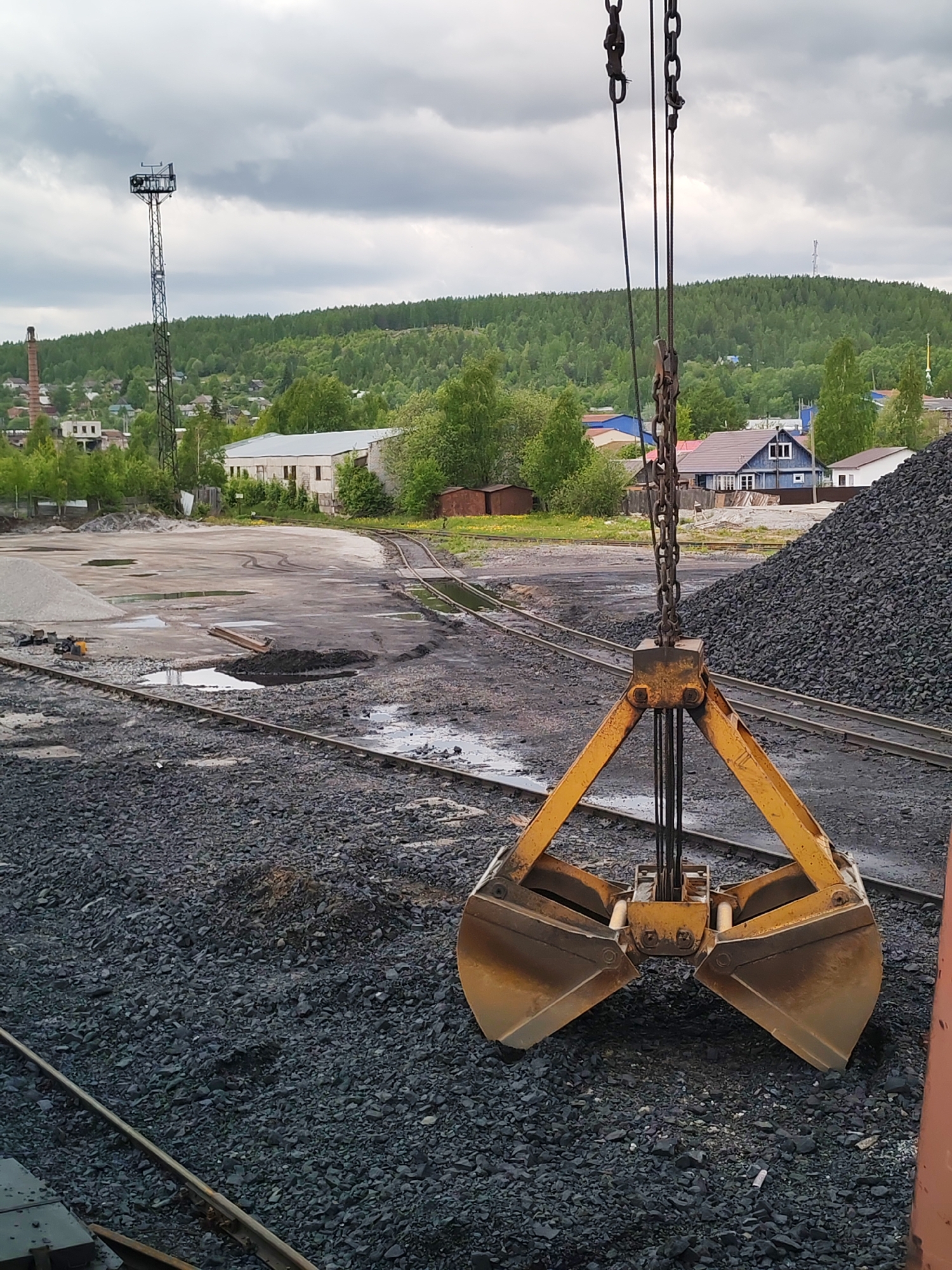The image depicts an industrial yard in Medvezhyegorsk, Russia, under a heavily overcast sky, suggesting a cloudy day or recent rain. In the foreground, a large, yellow-orange clamshell grab bucket, suspended by heavy chains and cables, hangs open over a ground covered in dark, angular bulk material, likely coal or crushed aggregate.
The immediate ground features more of this dark material, along with railway tracks running through puddles and muddy patches, indicating damp conditions. Further into the midground, a wide industrial area with multiple curving railway lines is visible, along with a pile of lighter grey aggregate. Distant, simple industrial buildings with concrete walls and metal roofs are partially obscured by greenery.
The background is dominated by a lush, green forested hill, characteristic of the region. Scattered among the trees and at the base of the hill are small residential houses, some with blue or brown roofs. A tall, black metal lattice tower, possibly for communication or floodlighting, stands prominently on the left, with another similar structure barely visible on the distant hill. A brick chimney can also be seen on the left. No people are visible, and there is no discernible text in the scene. The overall impression is one of an active, albeit somewhat rugged, industrial facility set against a natural backdrop.
