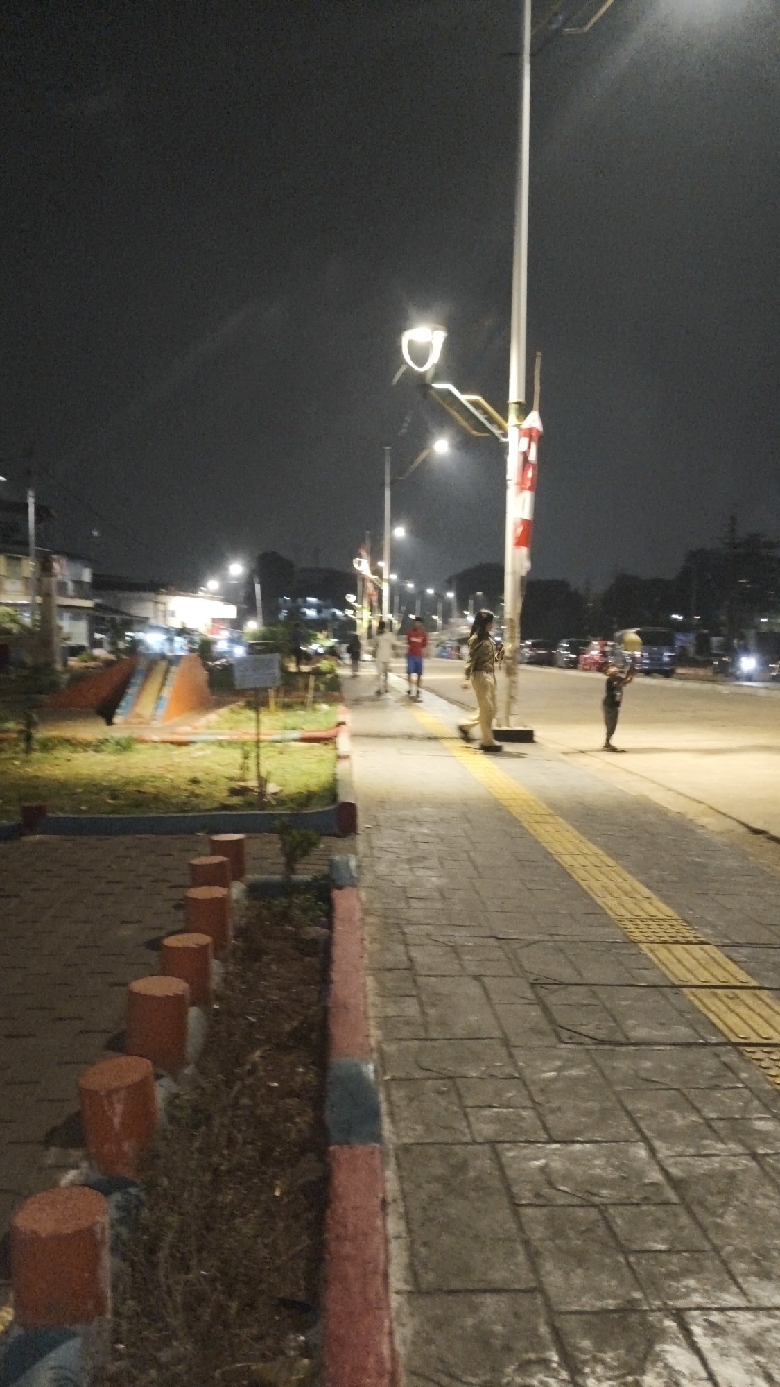 The image depicts a nighttime scene on a sidewalk adjacent to a road in South Jakarta, Indonesia. A streetlamp illuminates the area, casting a yellow glow on the tiled sidewalk, which features a tactile paving strip for the visually impaired. On the sidewalk, a woman in a uniform, possibly a security guard or traffic officer, stands near a street pole adorned with red and white stripes. To her right, a child is playing with a yellow ball. Further down the sidewalk, two other individuals are visible; one is wearing a red shirt and blue shorts, and another in a light-colored outfit. To the left of the sidewalk is a grassy area with a small playground structure featuring orange and blue slides. The background shows buildings and parked vehicles along the road, suggesting an urban environment. The overall atmosphere is dimly lit, characteristic of a city at night.