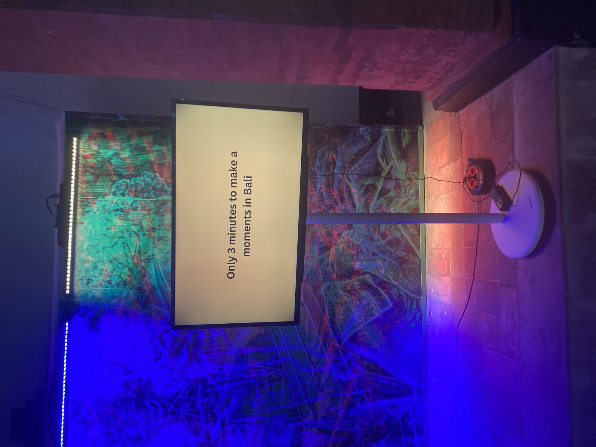 The image shows an indoor exhibition space in Kuta Selatan, Indonesia. A screen on a white stand displays the text "Only 3 minutes to make a moments in Bali." The background features abstract artwork in vibrant blue, green, and red hues. Lighting effects enhance the artistic atmosphere. A spotlight and electrical cables are visible near the floor. The surrounding architecture appears modern.